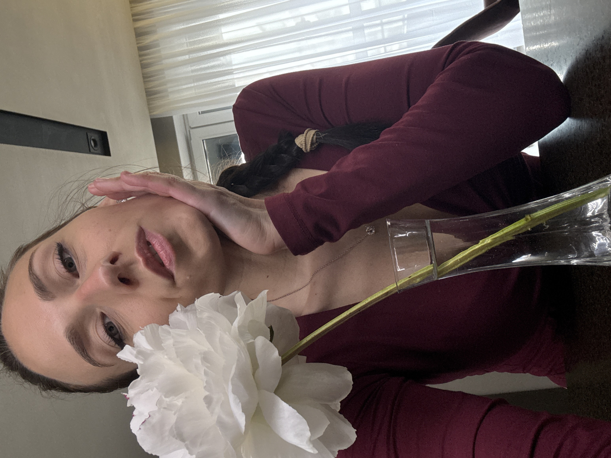 A woman with long dark hair, partially braided and secured with a tan hair tie, is holding a large white flower in front of her face. She is wearing a dark red, long-sleeved top. Her right hand is placed on her forehead. A slender, transparent glass vase containing a single green stem is positioned near her chest. A silver necklace with a small pendant is visible around her neck. The background features a window with white blinds and a dark gray fixture on the wall. The location is likely indoors. It cannot be determined from the image whether the location is in Saint Petersburg, Russia.