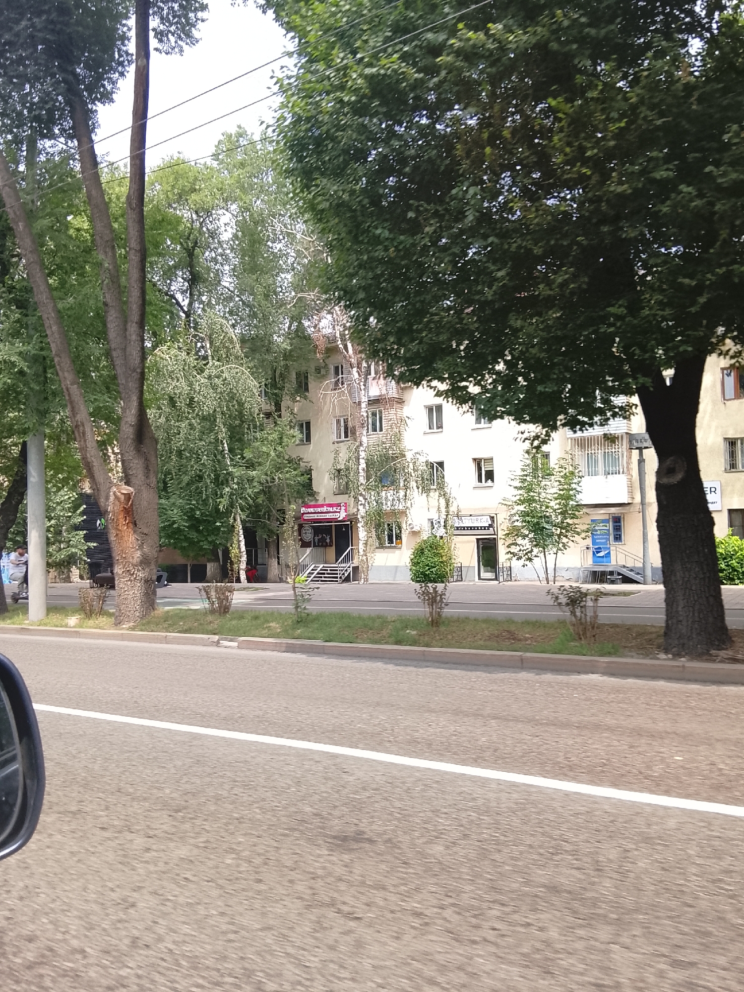 The image shows a street scene in Almaty, Kazakhstan, on a bright, clear day. The foreground is dominated by a paved road with a white line marking the lane. To the right, a car's side mirror is partially visible. The middle ground features a sidewalk lined with greenery, including trimmed bushes and trees. In the background, a multi-story building with several windows and balconies stands. A sign with Russian text and an image of a woman's face is visible on the building, possibly indicating a business. People are present, with one individual riding a scooter on the sidewalk and another person sitting near the building. The overall environment appears to be an urban setting with a mix of natural elements and man-made structures.
