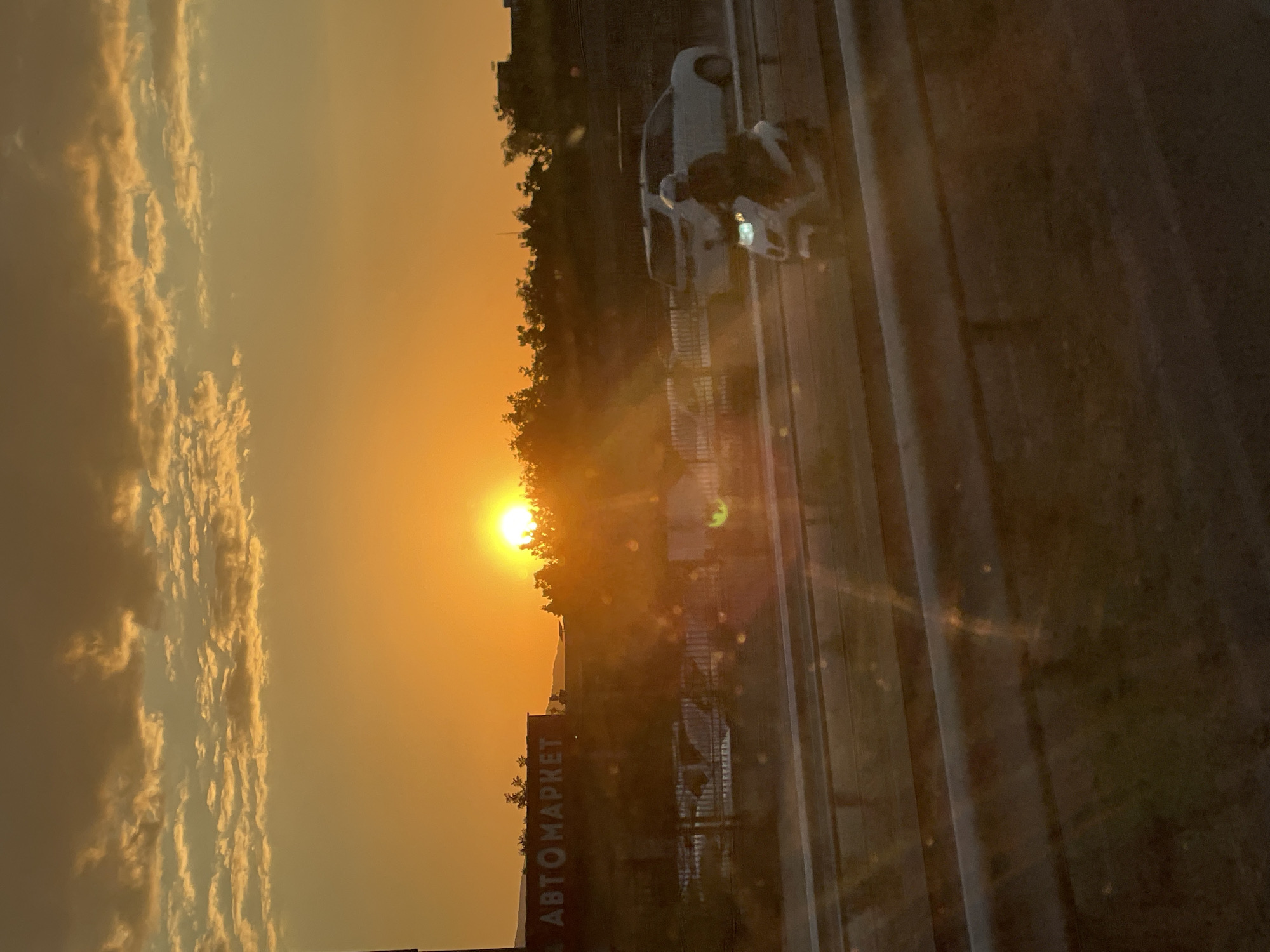 A sunset scene captures a highway and a building with the Cyrillic text "АВТОМАРКЕТ" visible. The sun is low on the horizon, casting a warm orange glow across the sky, illuminating scattered clouds. On the highway, a white scooter with a rider wearing a helmet is in motion, passing by parked cars, including a white SUV. The environment appears to be on the outskirts of Asenovgrad, Bulgaria, given the presence of the "АВТОМАРКЕТ" which translates to "Car Market." The overall atmosphere is serene, with the fading light creating a sense of tranquility.