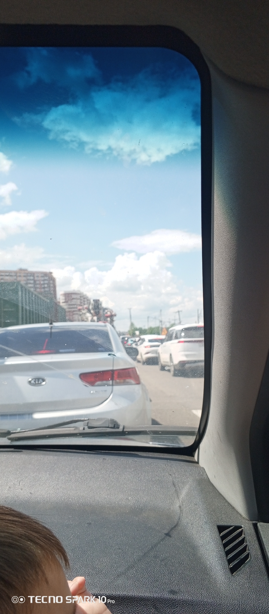 A view from inside a car in traffic. The sky is visible through the windshield. Other cars are in front, including a white Kia. The car appears to be in Belozernyi, Russia, based on the building architecture and vegetation visible. A child's head is visible in the lower left corner. The dashboard of the car is black. The sky is blue with white clouds. The camera model is TECNO SPARK 10 Pro.