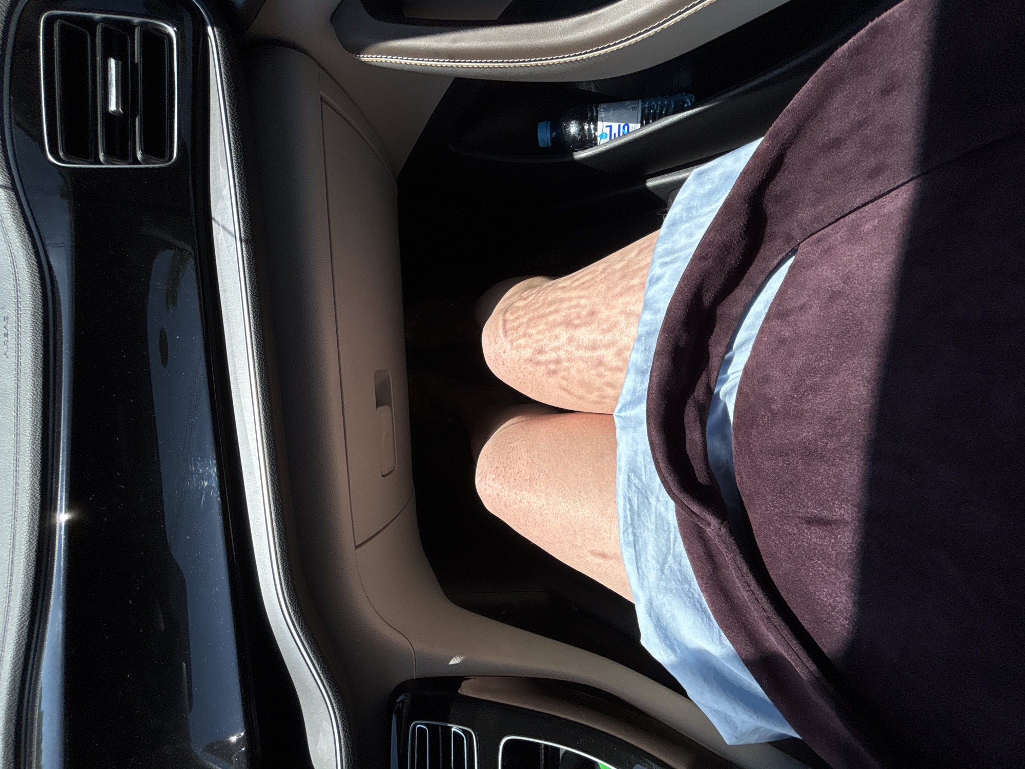 The image shows a first-person perspective of someone sitting in the passenger seat of a car. The interior of the car is visible, including the dashboard with air vents and a glove compartment. Sunlight is streaming in, casting patterns on the person's legs. The person is wearing a light blue garment and a dark purple garment. A bottle of water is visible in a cupholder. The scene is consistent with being inside a car in an urban environment during the daytime.