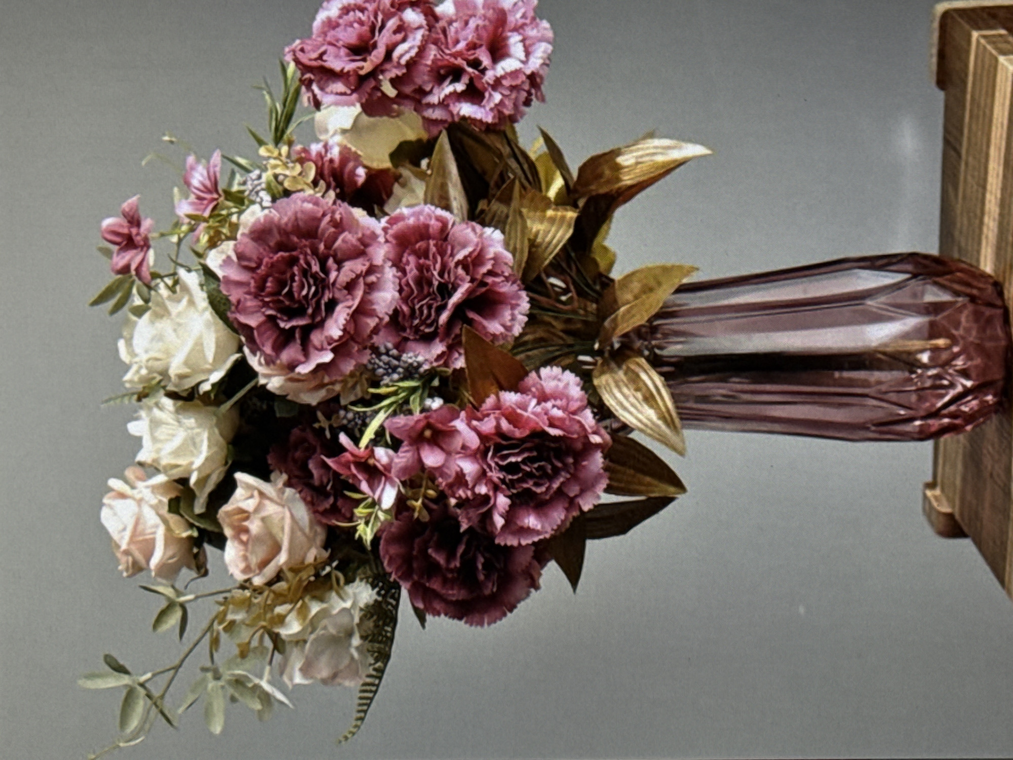 This is a studio photograph of a floral arrangement. The main subjects are a bouquet of artificial flowers and a decorative vase. The bouquet features a variety of flowers in shades of pink, purple, and cream, along with some dried, metallic-gold leaves and green foliage. Prominently featured are large, ruffled pink and purple carnation-like flowers, as well as smaller cream and light pink roses. The vase is made of a translucent, iridescent material, possibly glass or plastic, with a deep purple or burgundy hue and a faceted design. It is positioned to the right of the bouquet. Behind the floral arrangement, a light gray, neutral background is visible. To the far right, a portion of a wooden object, possibly part of a display stand or furniture, is present with vertical wood grain. The lighting is soft and diffused, suggesting a controlled studio environment rather than natural daylight. There are no people visible, nor any specific location cues to suggest the setting is Eyüpsultan, Turkey. The image is focused on the still life composition of the flowers and vase.