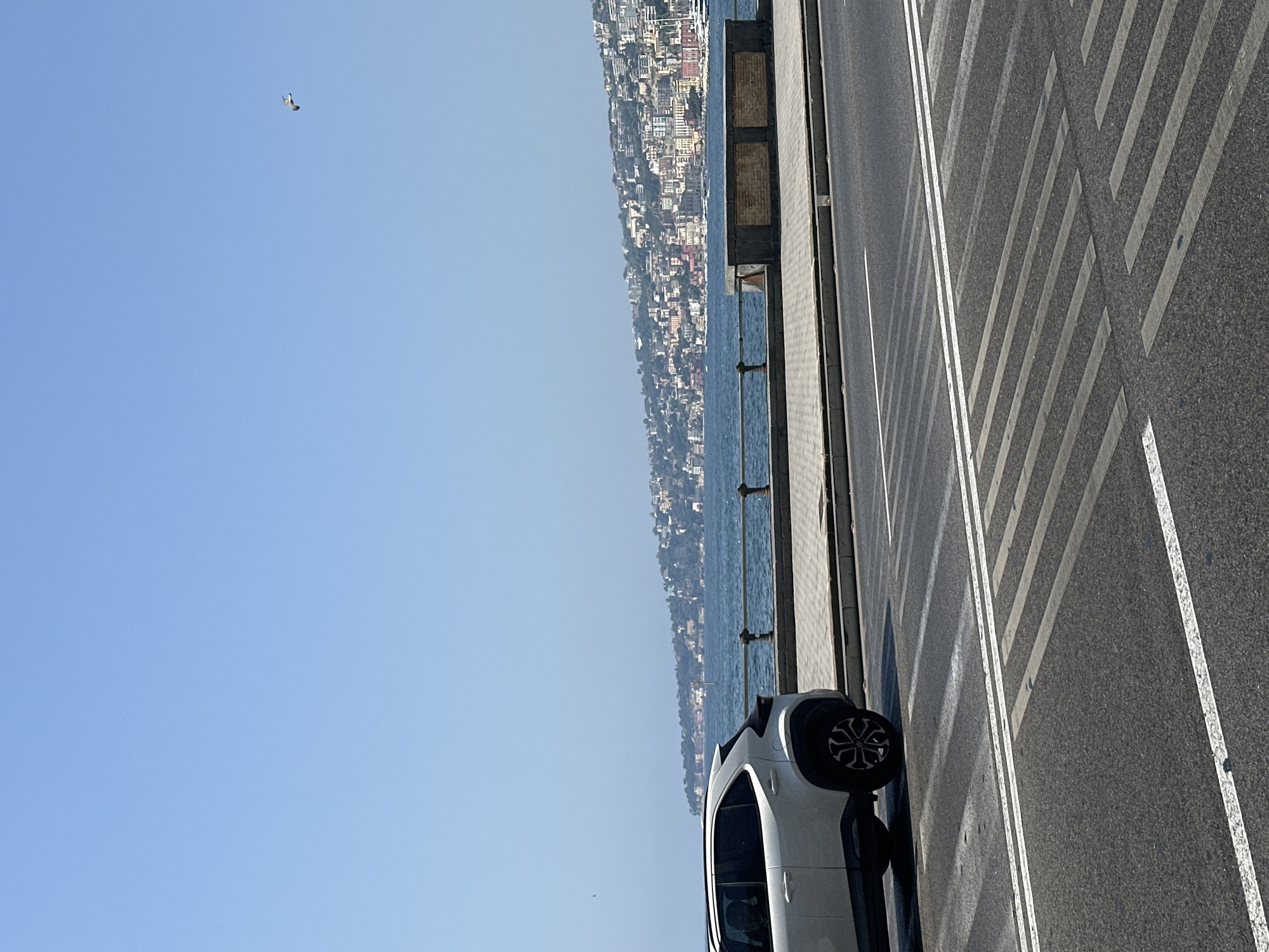 A white SUV is parked on the side of a road with a pedestrian crossing in the foreground. Behind the road, there is a walkway with a railing and a body of water. In the distance, a city skyline with buildings of various colors is visible along a hilly coastline. The sky is clear and blue with a bird flying in the upper right corner. The scene is likely in Naples, Italy, given the characteristic architecture and coastal setting.