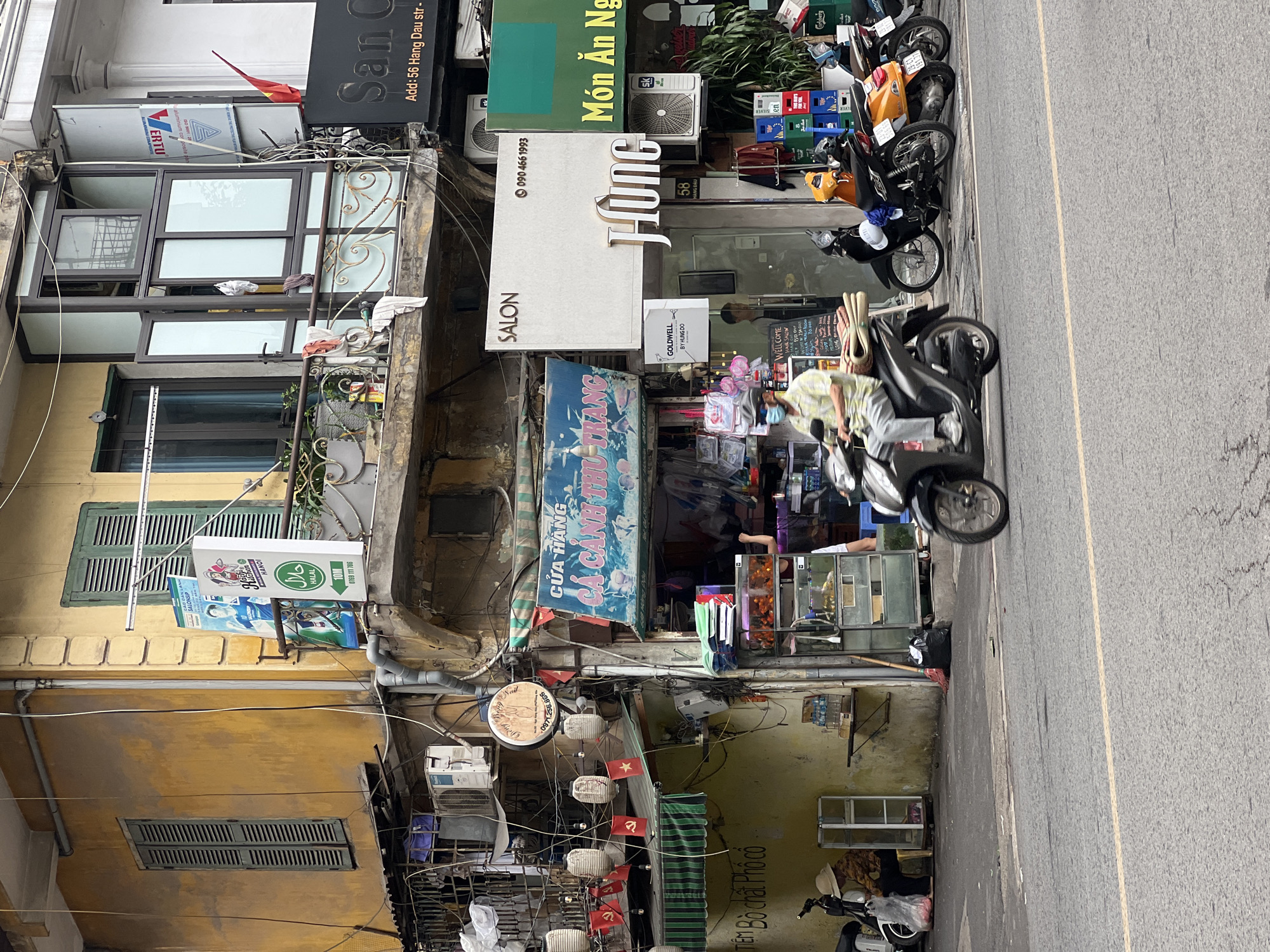 The image captures a street scene in Hanoi, Vietnam, showcasing a narrow, multi-story building facade with signs in Vietnamese. The building's exterior is a mix of weathered yellow stucco, old wooden shutters painted green, and modern glass and metal additions. On the street level, a store with a bright blue awning displays goods, possibly a market or shop. Several motorbikes are parked along the sidewalk, and one is in motion, ridden by a person wearing a mask and a light-colored shirt. The upper floors of the building reveal balconies with laundry hanging, and various signs advertising services like "SALON" and a phone number. A red Vietnamese flag is visible, adding to the national identity of the scene. The overall impression is of a busy, urban environment with a blend of traditional architecture and contemporary commerce.