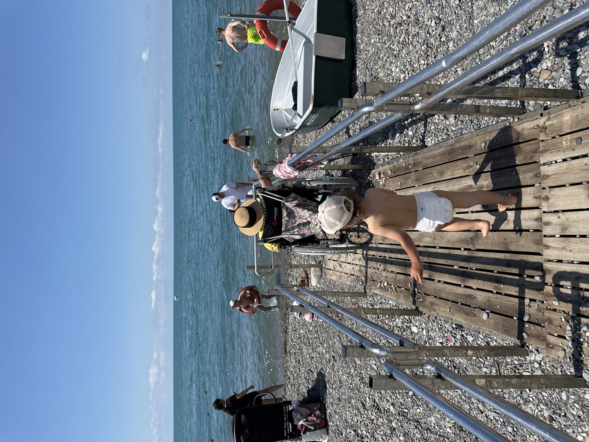 The image depicts a sunny day at a pebble beach, likely in Sochi, Russia, given the context. A wooden ramp with metal railings leads down to the water. In the foreground, a young child wearing a diaper walks away from the camera on the ramp. Behind the child, a person in a wheelchair, adorned with a straw hat, is positioned on the ramp. Further down the ramp, a small green boat is docked on the shore. In the background, several people are enjoying the water; some are swimming, while others stand near the shoreline. The sea is calm and blue, meeting a clear sky with a few scattered clouds. The overall atmosphere is one of a relaxed summer day at the beach.