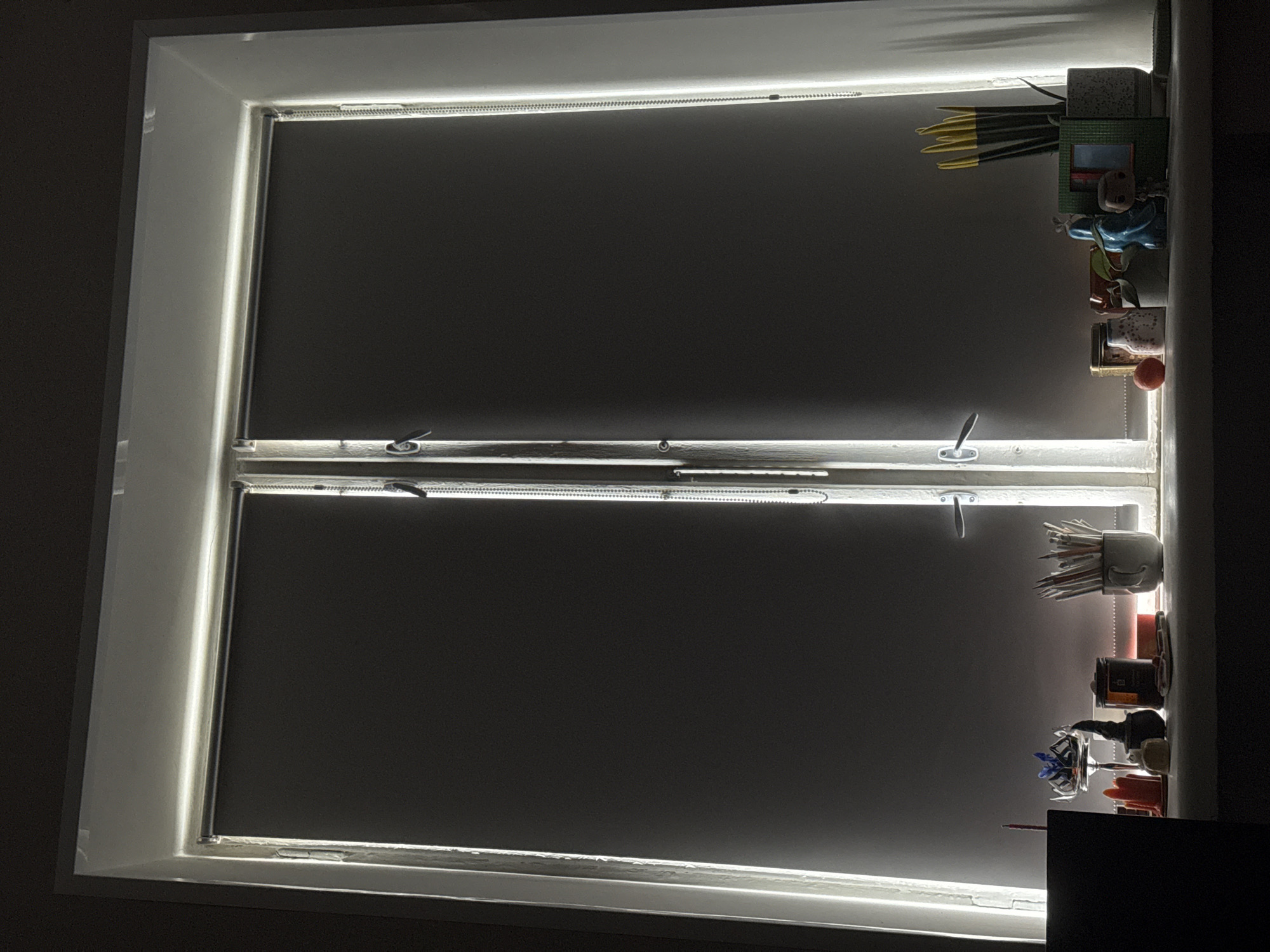 The image shows a double-hung window with dark-colored roller shades. The window is illuminated by LED strip lights on the frame, creating a bright perimeter. The window is partially open, with the lower sash raised. On the right side of the window frame, there's a collection of objects, including plants in pots, figurines, jars, and other small items. The overall impression is of a dimly lit room, with the window as the primary light source. The location is unknown, and there is no way to ascertain it from the image.
