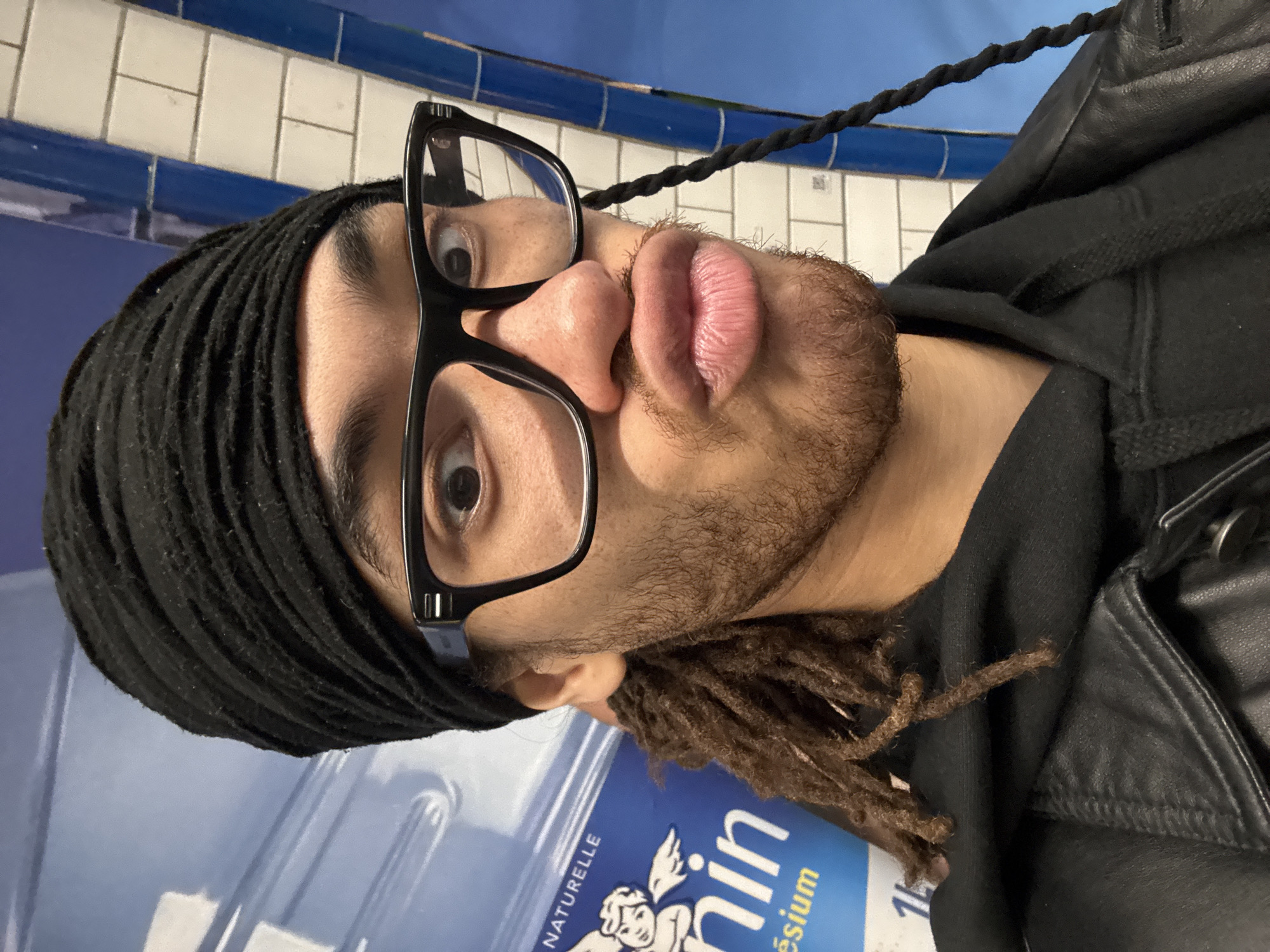 The image shows a person wearing a black head covering, black-framed glasses, a black hooded sweatshirt, and a black leather jacket. The person has light brown dreadlocks and a sparse beard. The person's face is positioned close to the camera, with their lips pursed.
The background features blue and white tiled walls characteristic of the Paris Métro. A blue and white bottle labeled "min" with the word "magnésium" is visible. The location is likely within a Paris Métro station.
