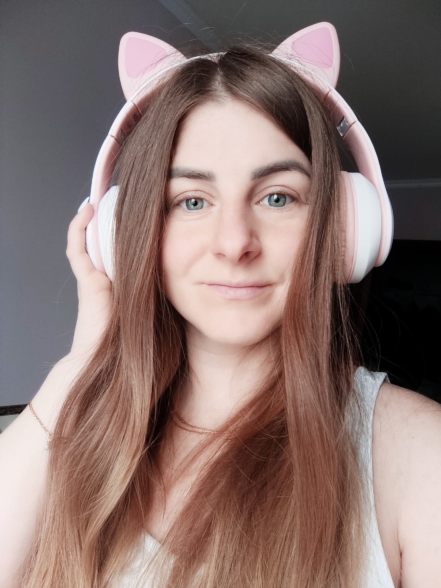 The image is a medium close-up portrait of a woman looking directly at the camera. She has long, straight brown hair that frames her face and extends over her shoulders. Her eyes are light blue-green, and she has a subtle, pleasant smile on her face.
The primary object she is wearing is a pair of pink and white over-ear headphones, distinguished by decorative pink cat ears attached to the top of the headband. Her left hand is lightly resting on the left ear cup of the headphones. Around her neck, a delicate gold chain necklace is visible, and a thin gold bracelet with a small charm can be seen on her right wrist, peeking from under her hair. She is wearing a light-colored top, possibly a tank top or camisole, with a visible strap on her right shoulder.
The scene is an indoor setting, with a dark, out-of-focus background that provides no specific details, suggesting a plain wall or shadowed area. The lighting is soft, illuminating her face evenly. The woman appears calm and approachable. There is no visible text in the image.