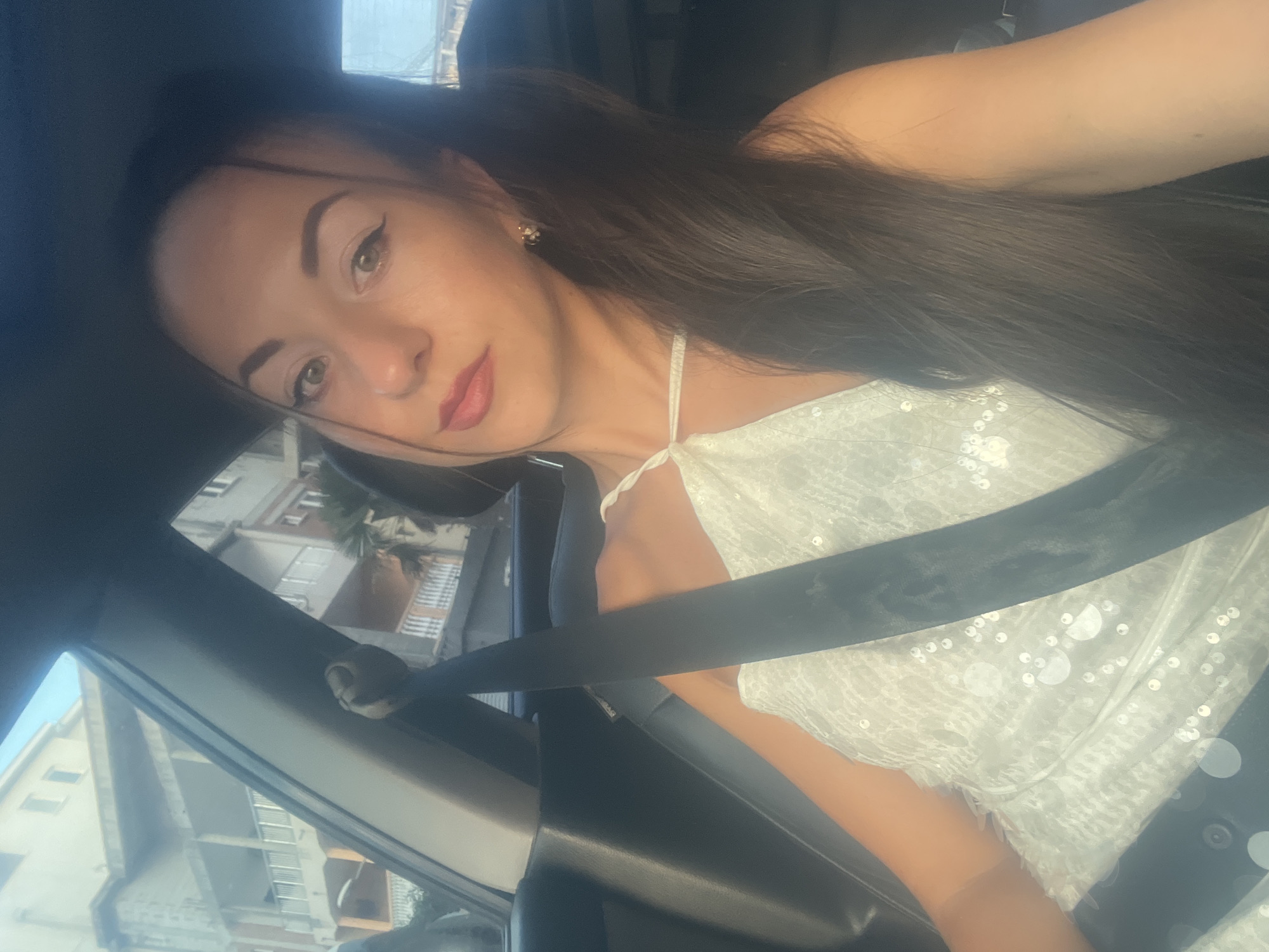 A woman is in the passenger seat of a car, wearing a white sequined dress and a black seatbelt. The woman has long, dark brown hair and is wearing makeup, including eyeliner and lipstick. She is looking at the camera. Outside the car, buildings and palm trees are visible. The scene appears to be in a sunny, urban environment.
