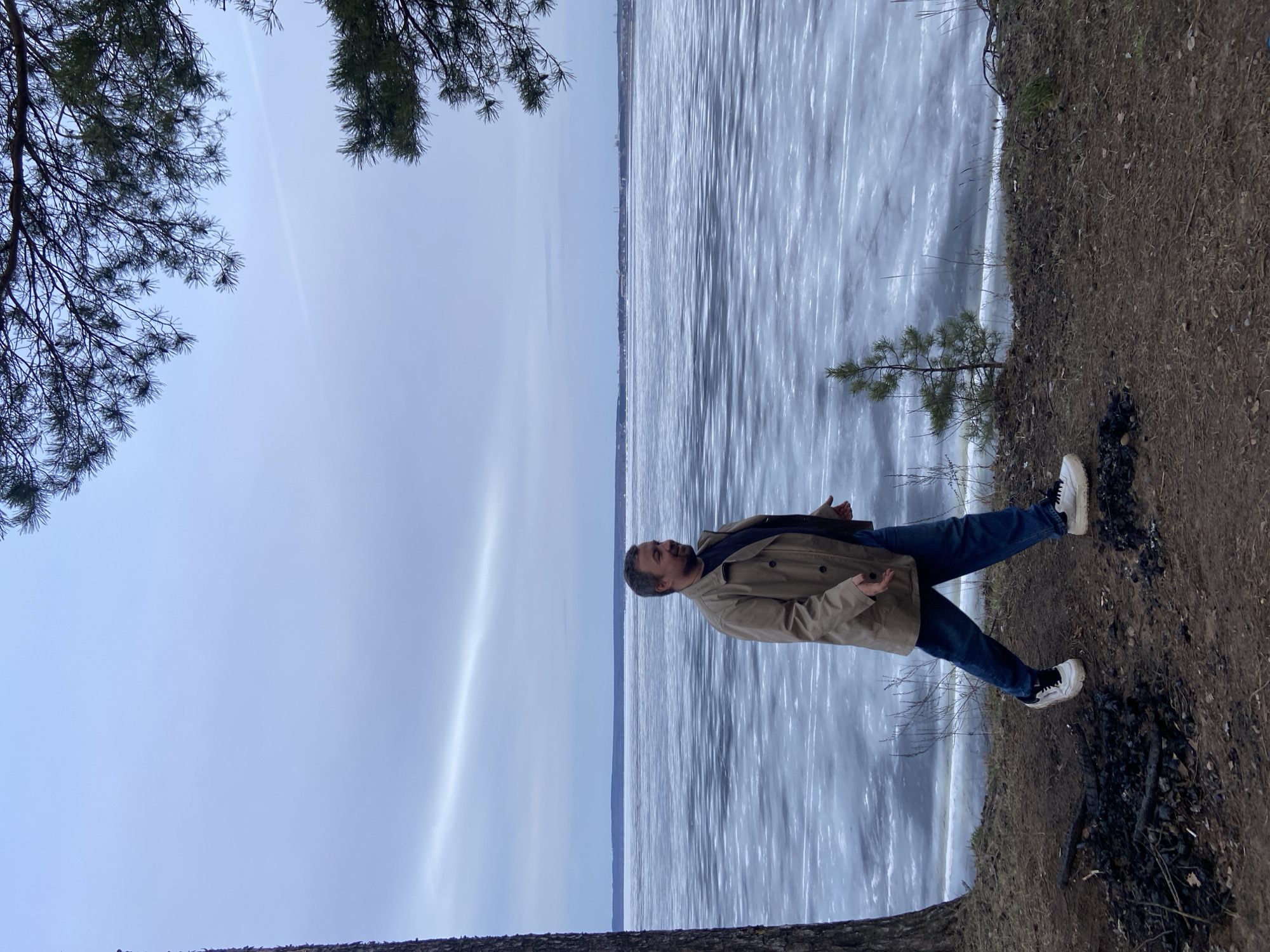 The image depicts a man standing near a body of water. The water's surface appears partially frozen or covered in ice. The man is wearing blue jeans, white shoes with black detailing, and a light brown coat. He is standing on the edge of a dirt bank, with some burnt remains of a fire nearby. Tree branches are visible in the corners of the image. The location is likely near Noviy, Russia, based on the context.