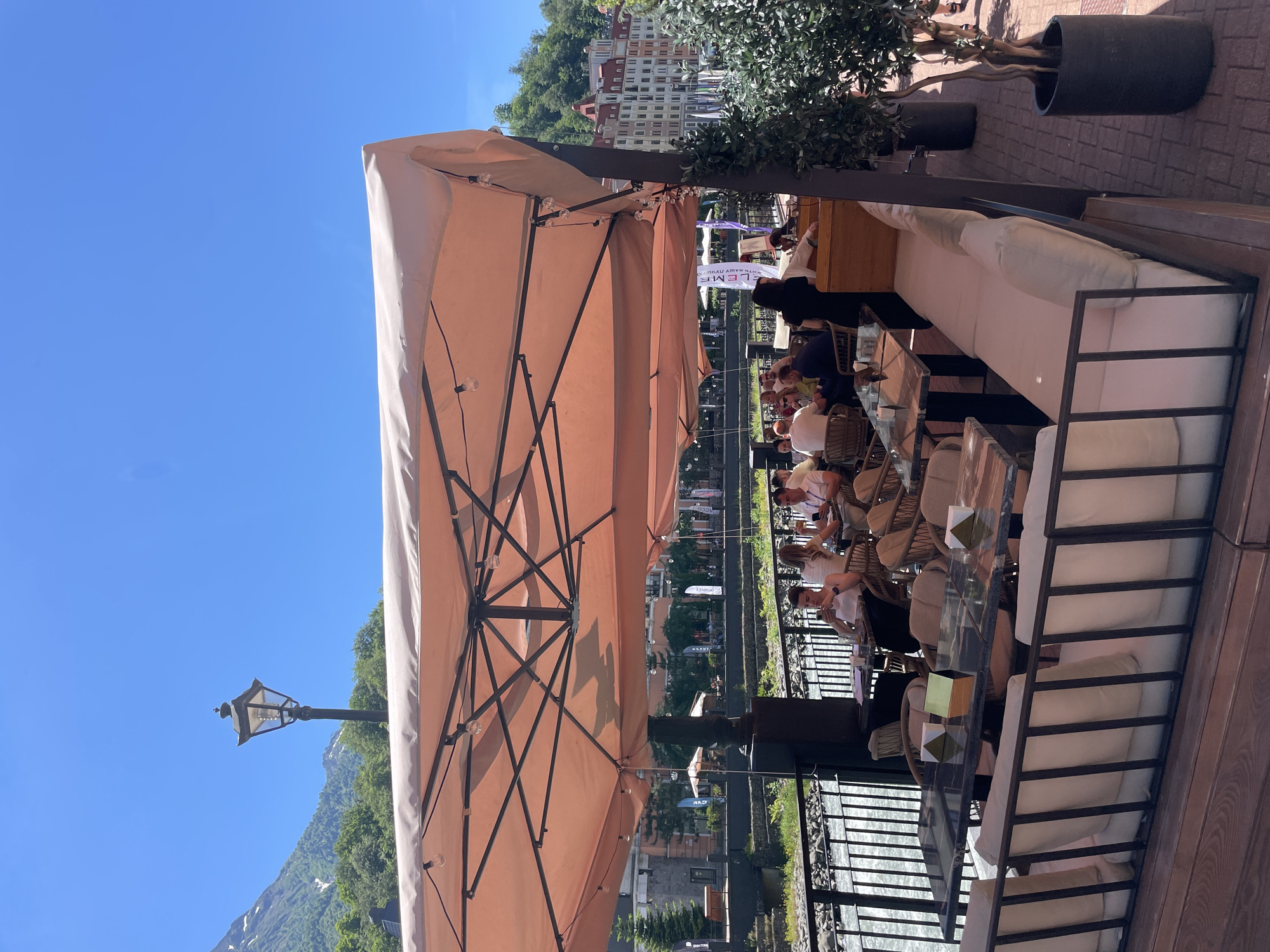 An outdoor cafe with a flowing river and mountains in the background. The cafe features a large, peach-colored umbrella covering multiple tables and seating areas. Several patrons are seated at tables, some dining, others looking at their phones. A server is visible behind a counter in the background. The cafe is situated in Estosadok, Russia, a resort town in the Krasnodar Krai region, known for its ski resorts and mountain scenery. The weather is clear and sunny.