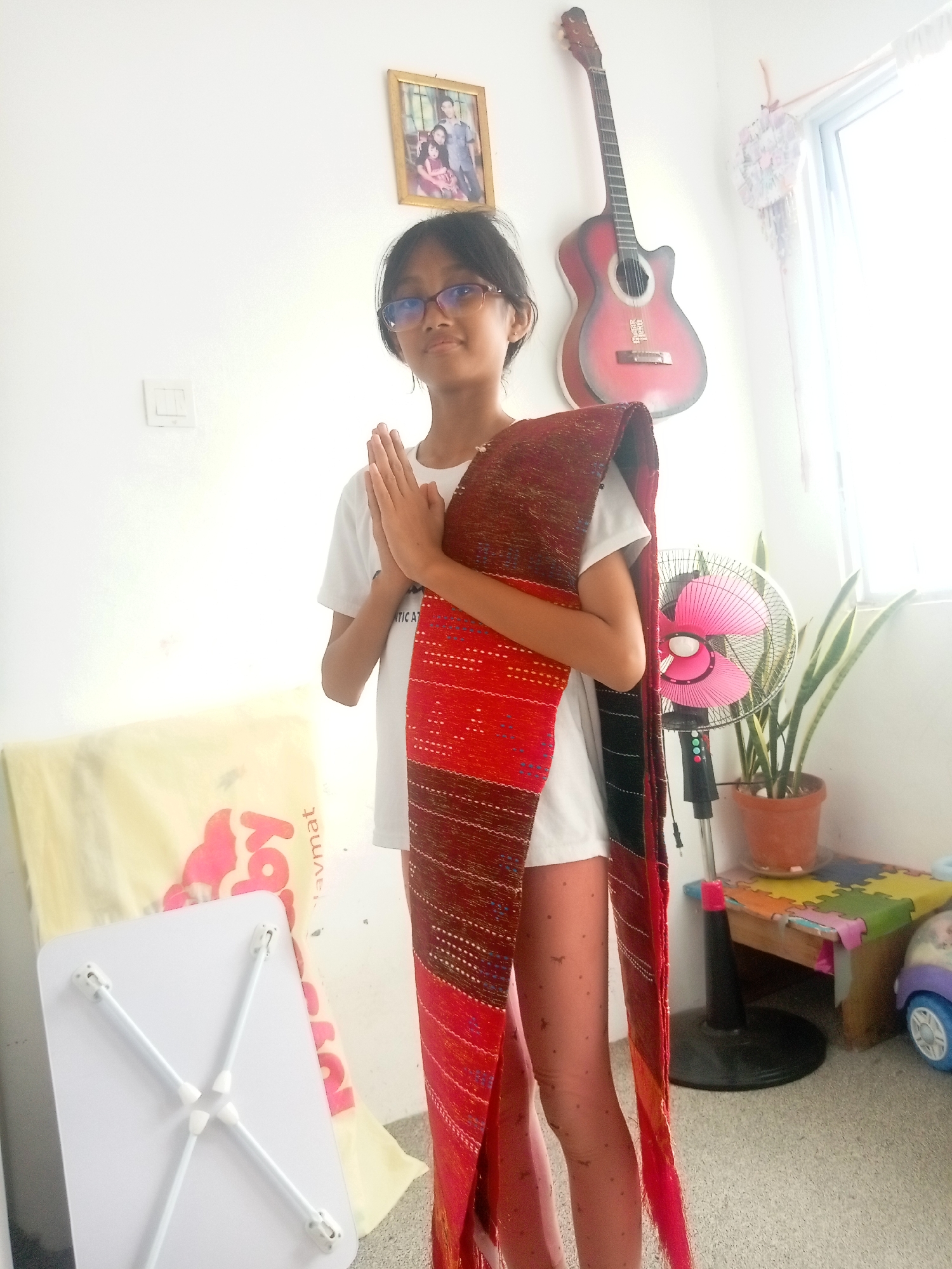 A young girl with glasses and short, dark hair stands in front of a white wall, holding her hands together in a prayer-like gesture. She is wearing a white t-shirt and patterned leggings, with a large, colorful woven fabric draped over her shoulders. To her left, a framed picture of a family hangs on the wall. Above and to her right, a red acoustic guitar is mounted on the wall. In the background, a pink electric fan is visible next to a potted snake plant. Further right, a child's play bench with colorful puzzle pieces sits near a toy car. A white foldable table is positioned in the lower left corner of the image. The scene appears to be indoors, in a room with neutral-colored walls and a tiled floor. The overall lighting suggests a bright, possibly daytime setting. The presence of the foldable table, the woven fabric, and the toy car hints at a domestic environment, possibly a living room or bedroom. The location is described as Batam, Indonesia.