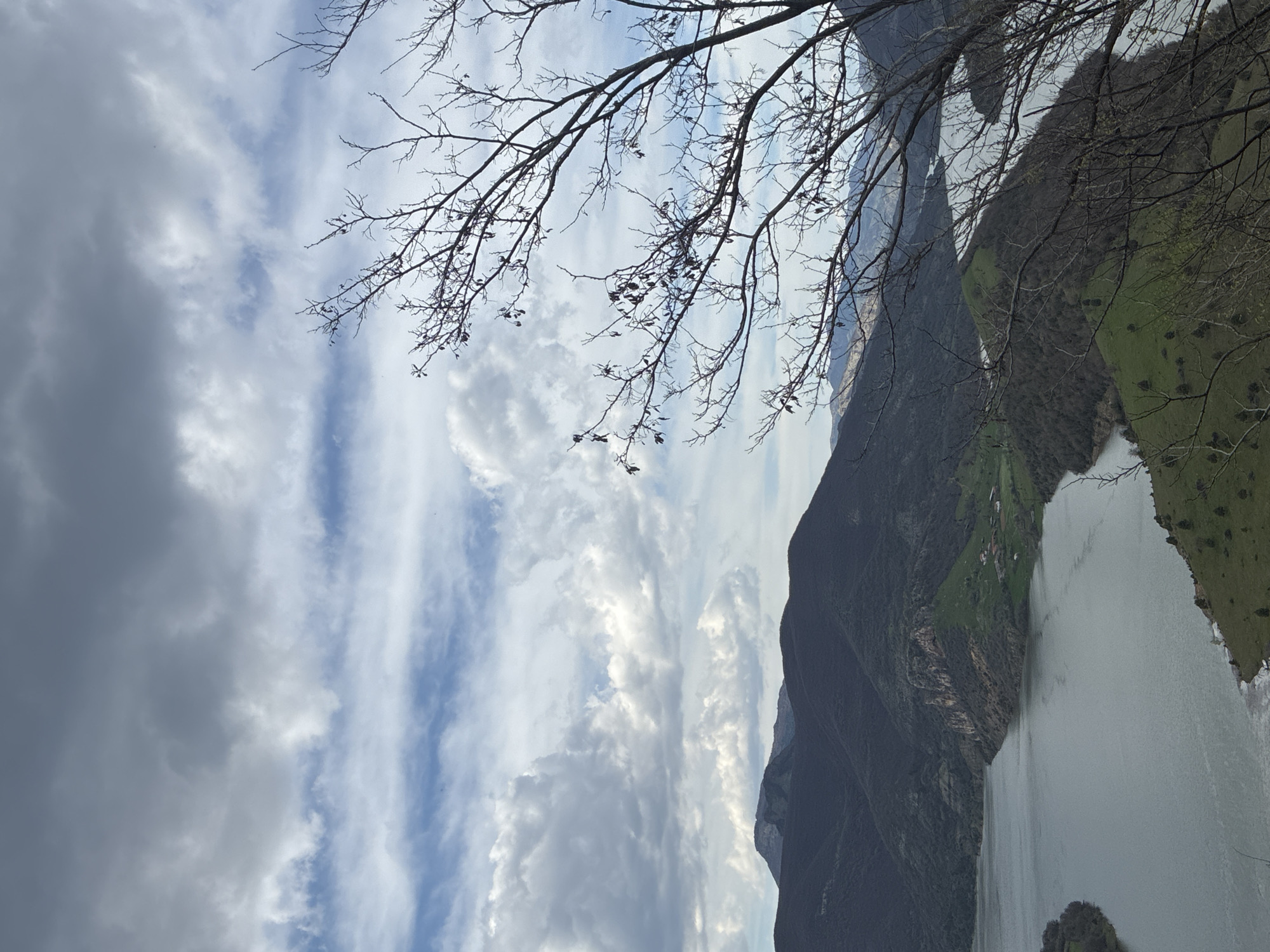 The image presents a landscape featuring a body of water, potentially a lake or river, flanked by steep, mountainous terrain. The mountainsides are partially covered in vegetation, with areas of exposed rock. The sky is filled with a mix of clouds, ranging from dark and ominous to bright and billowy, suggesting varying weather conditions. A bare tree branch extends across the top portion of the frame, adding a foreground element to the scene.
Location: Unknown, Unknown.