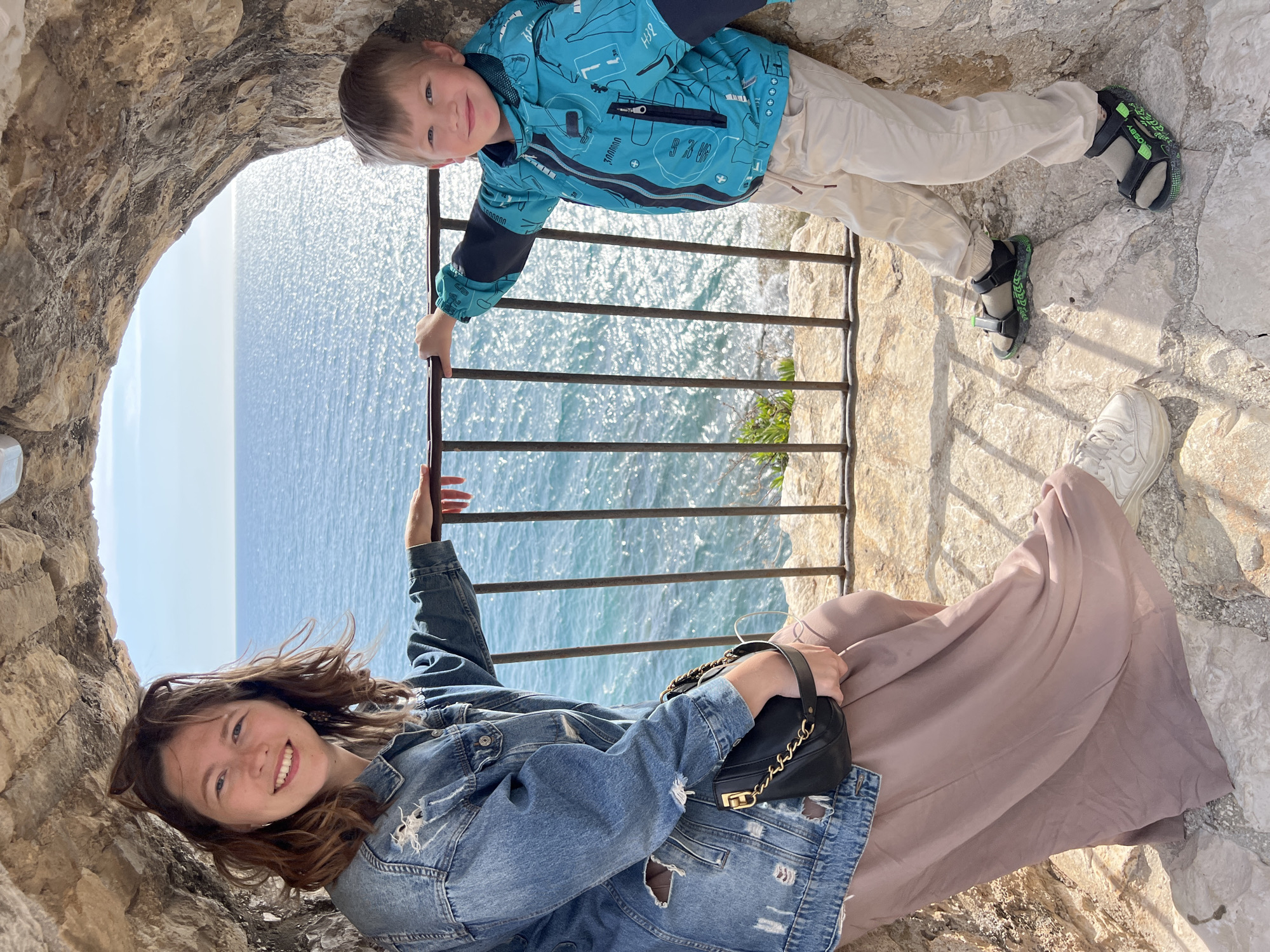 A medium indoor shot captures a woman and child posing within a stone structure overlooking the Adriatic Sea, with the location being the city of Ulcinj - Ulqin, Montenegro.
A woman with curly brown hair, wearing a denim jacket with rips, and a long mauve-colored skirt, is shown from the torso up. She is smiling and holding a black handbag with a gold chain.
Next to her, a fair-skinned boy with blond hair is wearing a blue jacket with a graphic pattern and khaki pants. He is also smiling. He has on green and black sandals.
The pair are positioned within a stone archway, a stone wall is visible to the left, and the ocean and sky are seen through an opening. A black metal railing separates the subjects from the sea. The flooring is made up of rough stone slabs.
The lighting is natural. The composition of the image is slightly tilted.