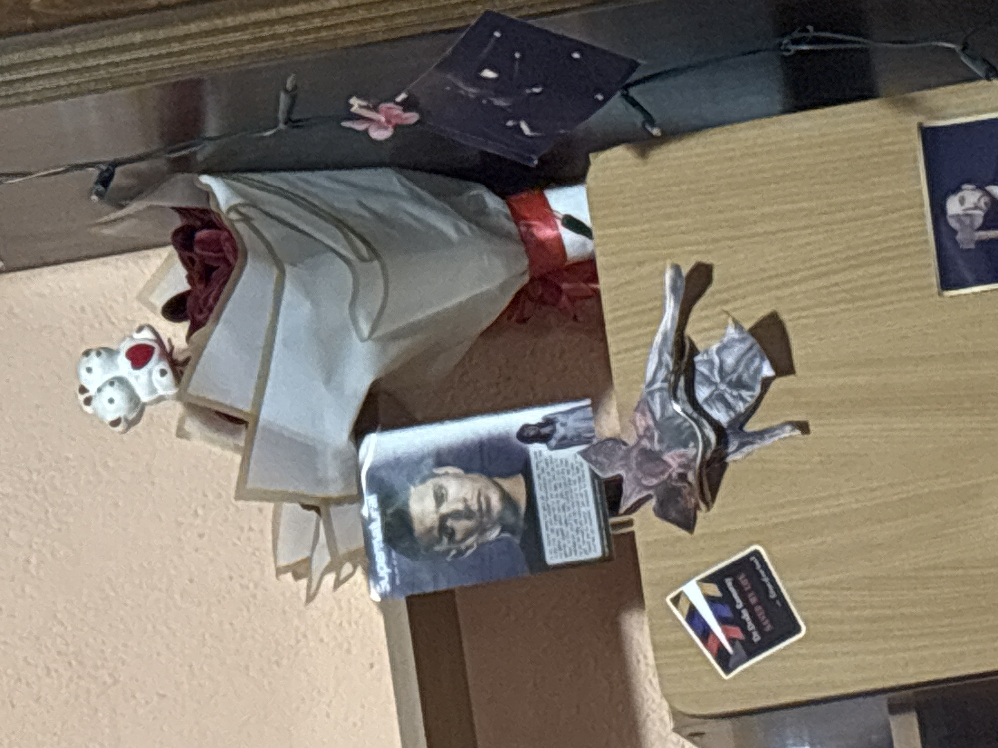The image shows a cluttered table surface. A bouquet of red roses wrapped in white paper with a red ribbon lies on the table. A card with a photograph of a man is placed nearby, along with a silver decorative flower. A small sticker with the text "The Duke Company SAVED MY LIFE - Good for you!" is also visible. The scene is lit by string lights, some of which are visible in the background. The location is likely indoors but cannot be confirmed to be in Garmen, Bulgaria without further information.