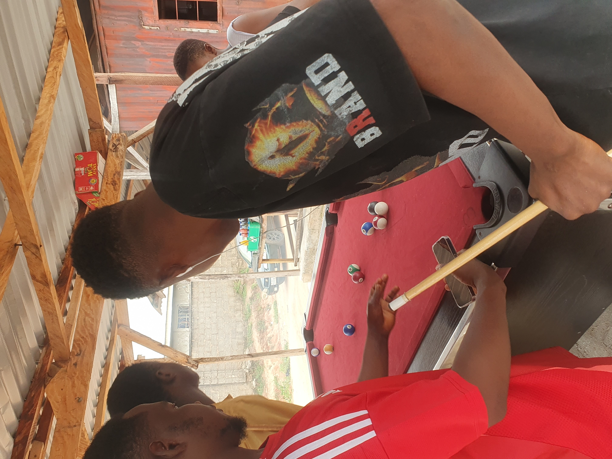 An outdoor, informal gathering occurs at a pool table in Lokoja, Nigeria, under a wooden and corrugated metal roof structure. Three individuals are partially visible, with two in the foreground. A person on the left wears a red shirt with white stripes and is about to play pool, holding a cue stick. To their right, another individual in a black t-shirt with a graphic design and the word "BRAND" is also present. A third person is visible further back. The pool table has several colored balls scattered across its red felt surface. The surrounding environment includes a dirt ground, a road with a parked car in the background, and brick buildings. There are also some crates and a red metal corrugated structure in the background, suggesting a makeshift or open-air establishment. The overall scene depicts a casual leisure activity in an urban, possibly semi-developed area.