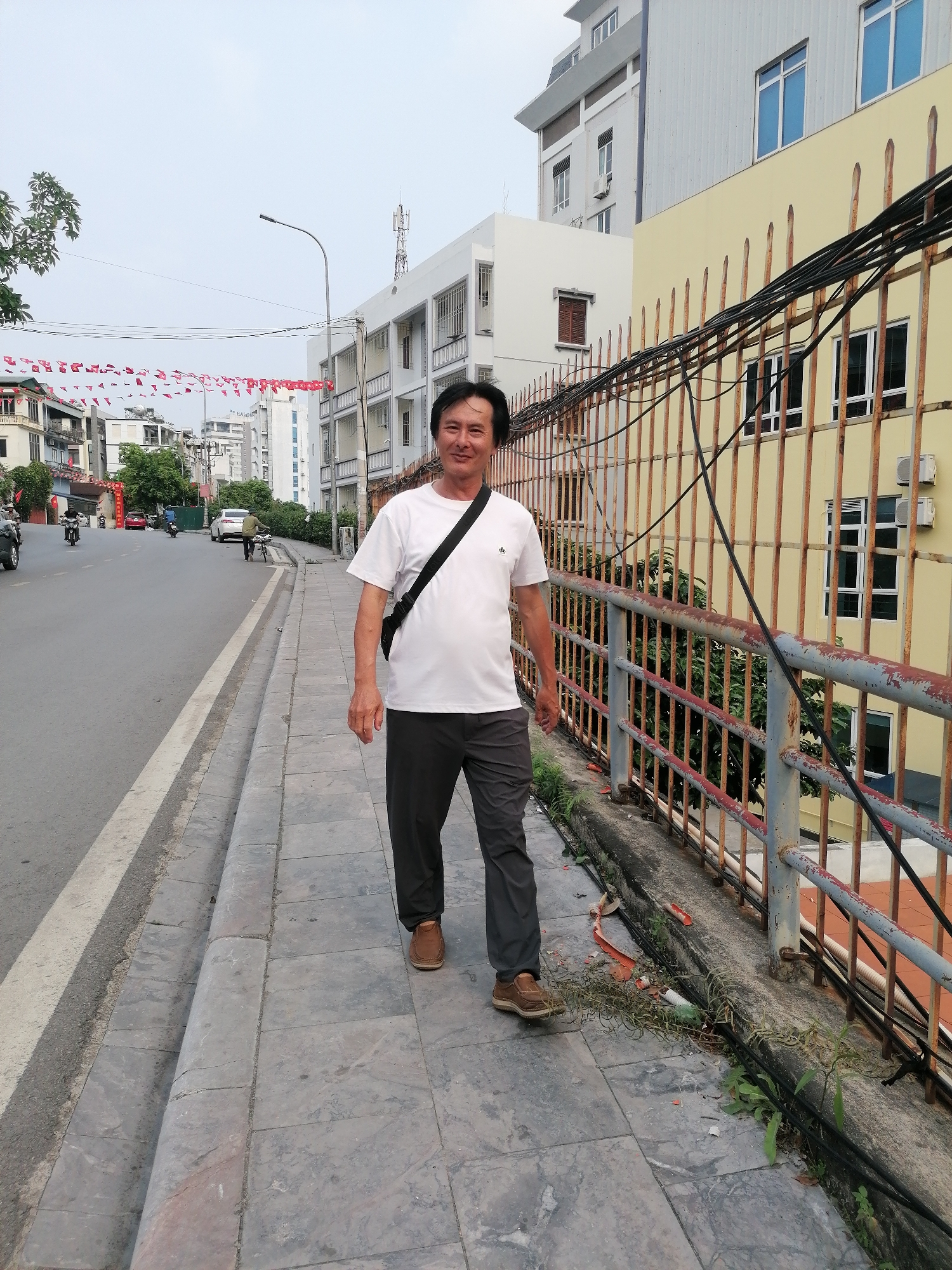 An Asian man in his 40s or 50s is captured walking towards the camera on a paved sidewalk in an urban setting, identified as Hong Gai Ward, Vietnam. He is smiling pleasantly and wears a white short-sleeved t-shirt with a small "db" logo on the left chest, dark gray trousers, and brown shoes. A black messenger bag is slung across his body.
To the man's right, a tall, rusted metal fence runs alongside the sidewalk, heavily draped with black electrical wires. Beyond the fence, a light yellow building with several windows and external air conditioning units is visible, backed by a taller white building. The sidewalk itself shows some wear, with small patches of greenery and debris near the fence.
To his left, an asphalt road with a white lane marking extends into the distance. Motorbikes and cars, along with other pedestrians, can be seen on the street, indicating moderate traffic. Red triangular flags, likely celebratory banners, are strung overhead across the street. The sky is bright and appears overcast, suggesting either a cloudy day or early morning/late afternoon light. The scene depicts a typical busy street environment in Vietnam, characterized by a mix of buildings, infrastructure, and local decorations.