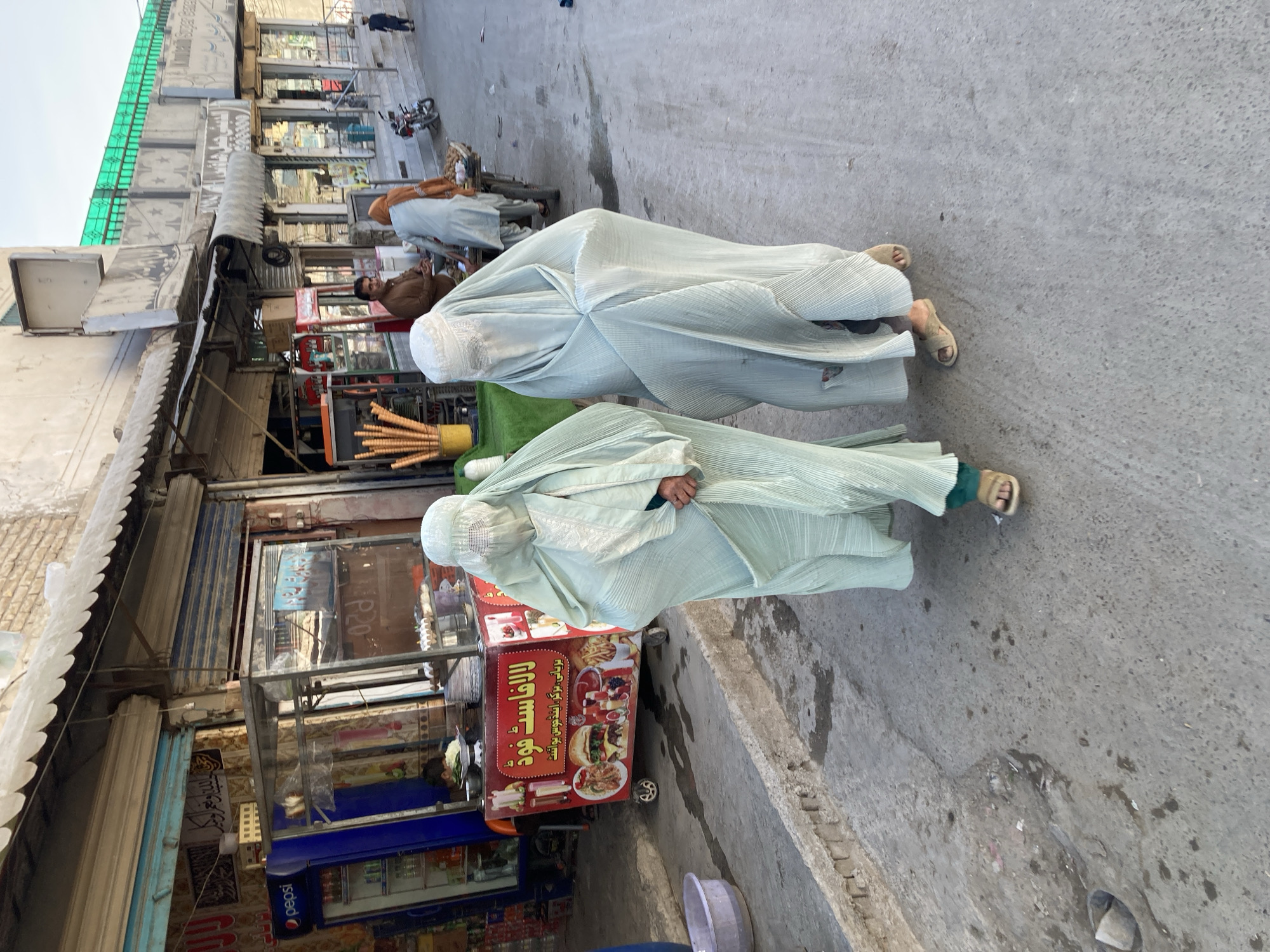 Two women completely covered in light green garments are standing on a paved city street in Quetta City Tehsil, Pakistan, near storefronts and a food cart. The women are wearing flat sandals. One woman stands adjacent to a red food cart. The cart displays a variety of food items in Arabic script and images of burgers, fries, and other meals. The storefronts behind the cart include shops selling various goods, including Pepsi products. Behind the women, there's a man sitting near a motorcycle. Other people are walking in the background. The sky is visible.