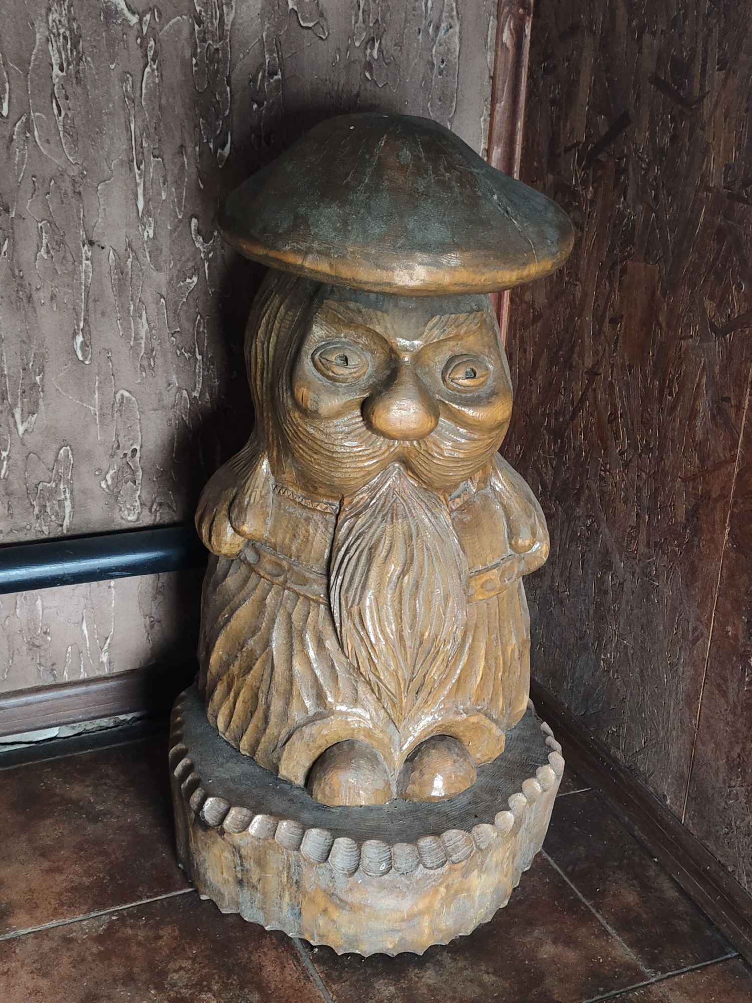 A wooden carved figure of a gnome-like character stands prominently in a corner. The gnome wears a hat and has a long beard. It's placed on a carved wooden base with rounded edges. The background consists of a textured wall on one side and wooden paneling on the other, suggesting an indoor setting. The figure is illuminated from an unseen light source. The exact location is unknown.