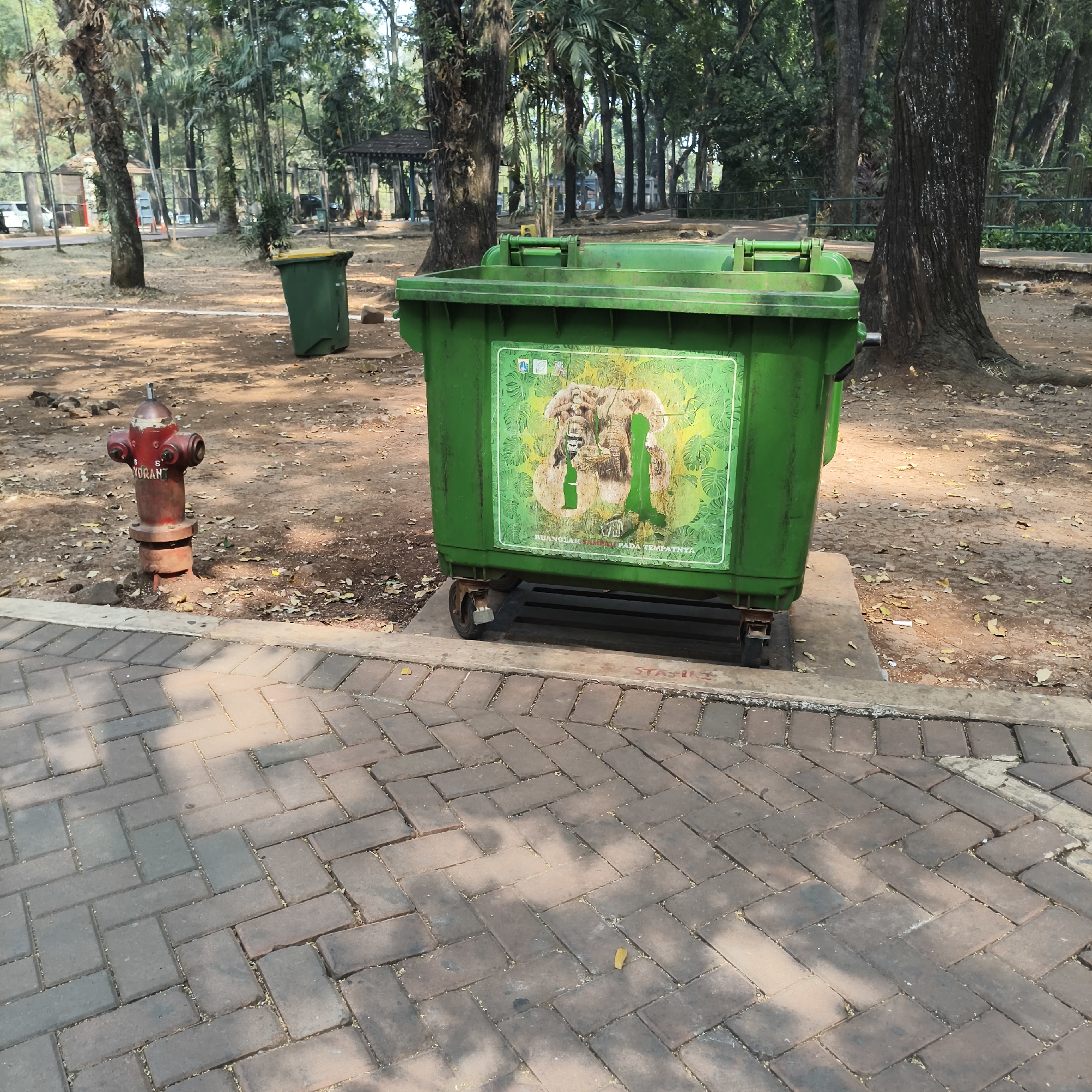 A bright green, large capacity trash bin with wheels sits on a concrete slab in a park. The bin has a graphic depicting elephants and foliage. Next to the bin is a red fire hydrant. The ground around the bin and hydrant is covered in dirt and fallen leaves. A paved walkway made of red brick with a herringbone pattern leads away from the bin and hydrant. In the background, there are numerous trees, some with dense foliage, and a wire fence. A smaller green trash bin with a yellow lid is visible further in the background. This scene appears to be located in Kebun Binatang Ragunan (Ragunan Zoo) in South Jakarta, Indonesia, given the presence of the graphic of elephants on the trash bin and the general park-like environment common in Indonesian cities.