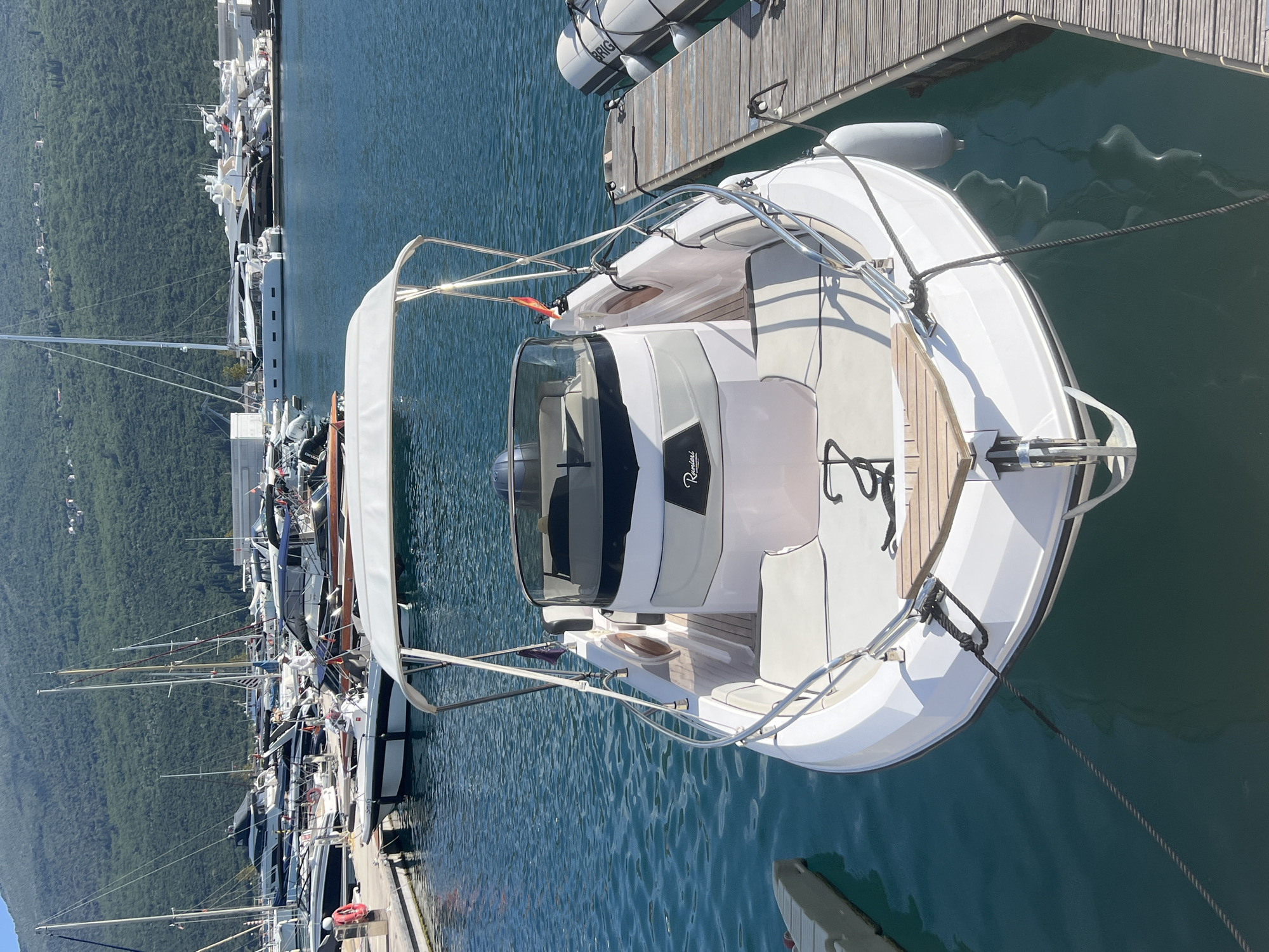 The image captures a vibrant marina scene in Đenovići, Montenegro, bathed in bright daylight under a clear sky. In the foreground, a sleek white and grey motorboat is prominently docked. It features a white canvas canopy, a grey and white interior, a dark windshield, and a light wooden deck at the bow with a stylized anchor illustration. The brand "Ranieri" is visible on its side near the console.
The main boat is secured to a wooden pier by mooring ropes. Along the pier, numerous other boats are docked, including large luxury yachts, several sailboats with tall masts, and other motorboats, indicating a bustling harbor. A white inflatable fender attached to the dock clearly shows the brand "BRIG."
In the background, a vast, densely forested mountain range dominates the horizon, providing a dramatic natural backdrop to the man-made harbor. The water in the marina is a clear, deep blue, with visible ripples reflecting the sunlight. A few indistinct figures can be seen on some of the distant boats and docks, but no specific activity is highlighted. The scene conveys a sense of tranquility and leisure in a picturesque coastal setting, typical of a busy holiday destination.