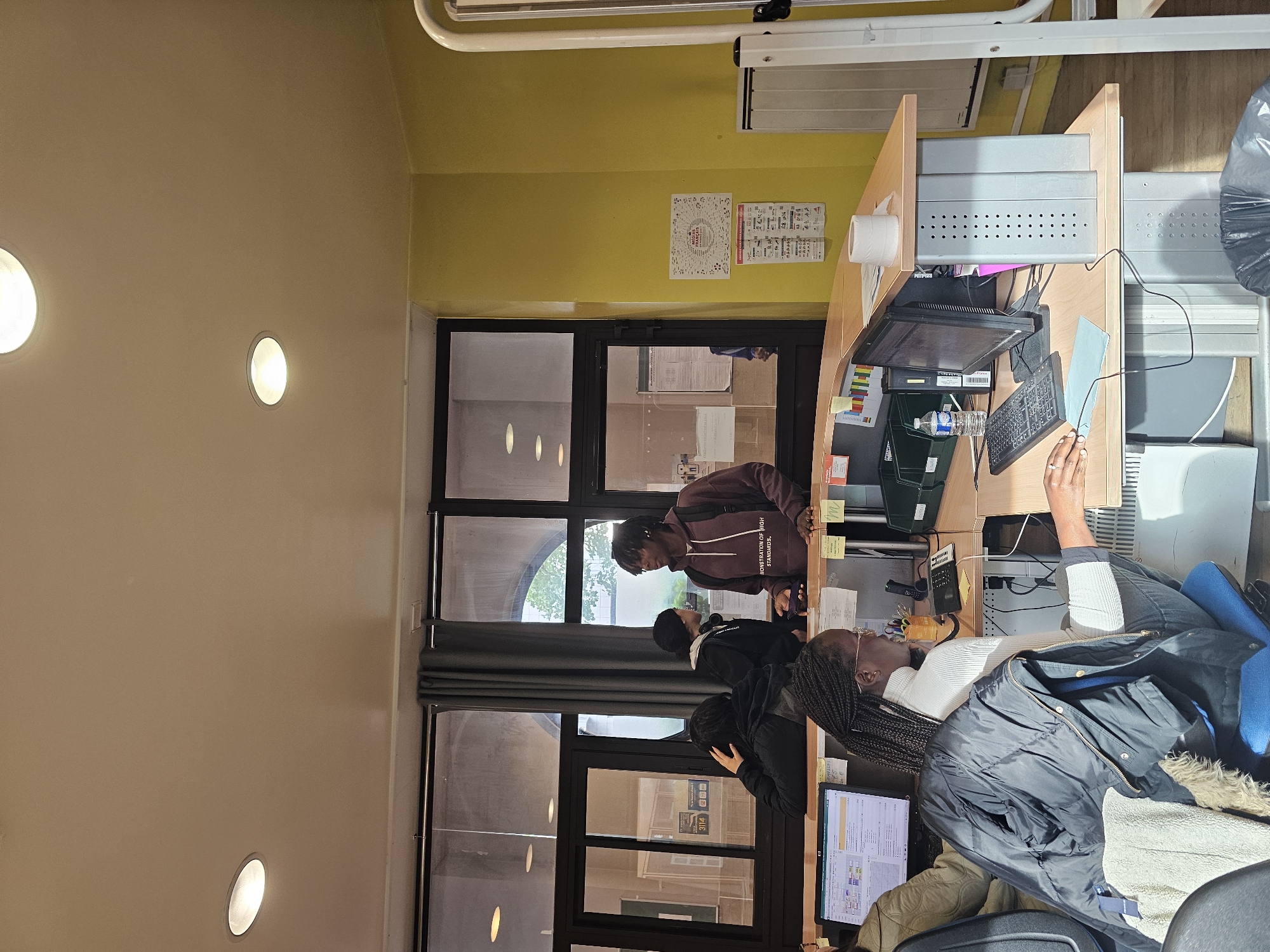 The image depicts a group of people, primarily students, interacting at a reception desk, likely in an academic building or a similar institution in Paris, France. The location is suggested by architectural elements and the visible urban landscape through the windows, possibly indicating a setting near a park or a building with historical significance.
The scene includes at least four individuals of African descent. One individual stands at the desk, possibly seeking information or assistance, while others are either waiting or engaged in conversation near the reception area. The desk itself features computers, monitors, and administrative supplies.
The interior environment suggests a modern space, characterized by yellow walls and minimalist lighting, contrasting with the dark metal framing the doors and windows.