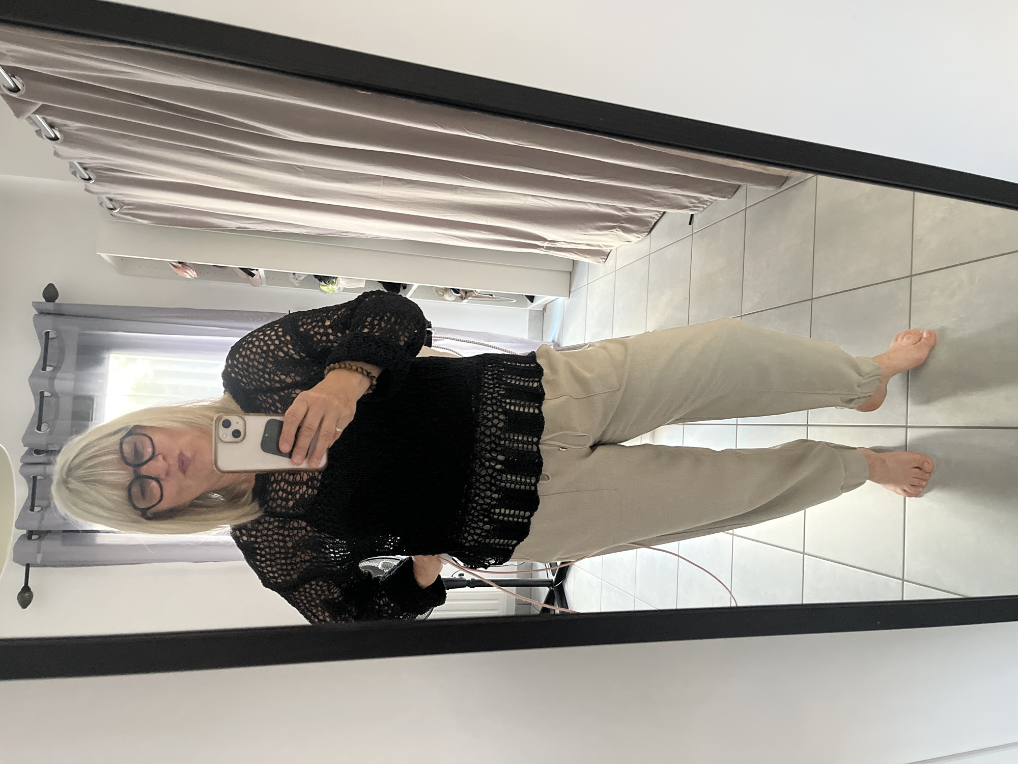 A woman is taking a selfie in a mirror. She has short blonde hair, glasses, and is wearing a black crocheted top and tan pants. She is barefoot and holding a white iPhone. The background includes a curtain, a shelf with items on it, and tiled flooring. The image was likely taken indoors in a residential setting in Chambœuf, France.