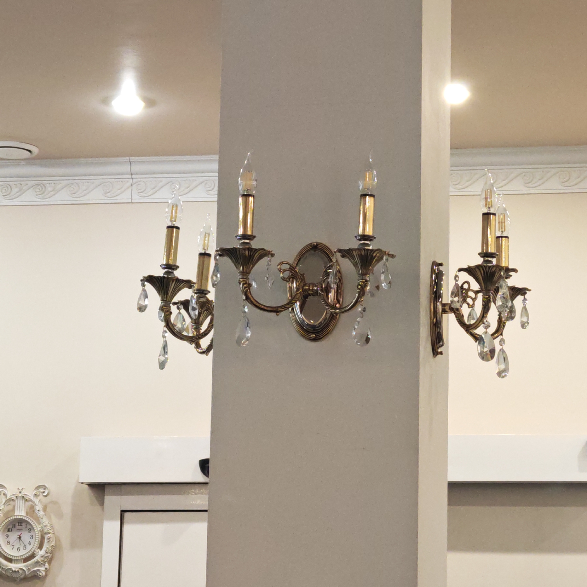 This media file presents an interior scene in Kladovitsy, Russia, focusing on decorative lighting and architectural details. No people are visible.
The central feature is a prominent, light-brown vertical partition or column. Mounted on this column are two ornate, antique-style wall sconces made of brass or bronze. Each sconce holds two 'candle' style light bulbs, which are illuminated, and features multiple hanging crystal drops that refract light. A third, similar sconce is partially visible on the far left wall.
The walls are painted a light, neutral beige or cream color. White decorative crown molding with a repeating scroll pattern runs along the top perimeter of the walls. A bright, recessed ceiling light is visible on the ceiling to the left, with another implied or reflected on the right.
In the lower-left corner, a white, ornately framed wall clock with Roman numerals displays the time as approximately 8:07. Below the crown molding, a simple white shelf or ledge extends, and part of a white door frame or cabinet is seen in the bottom left. The overall impression is one of classic or traditional interior design, with a focus on decorative elements and warm artificial lighting, suggesting either morning or evening.