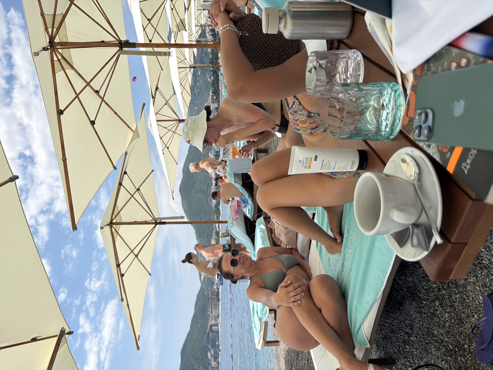 The image depicts a sunny day at a beach resort, likely in Budva, Montenegro, with several people relaxing under umbrellas. In the foreground, a woman in a green bikini and sunglasses is seated on a towel, looking towards the camera with a relaxed expression. She has her hands clasped in her lap. To her left, a white cup sits on a saucer, next to a metallic object and a dark brown wooden table.
In the background, multiple cream-colored beach umbrellas are set up, casting shadows on the pebble beach and the turquoise water. Other individuals are visible lounging on chairs or walking along the shore. A red paraglider can be seen in the sky, adding a sense of leisure and recreation to the scene. The weather appears to be clear and warm, typical for a beach destination.
Various objects are scattered around, including glasses, a bottle of sunscreen with the text "HELIOCARE Advanced Gel SPF 50" clearly visible, and a green iPhone. The overall atmosphere is one of a leisurely vacation day at the seaside. The mountainous landscape with buildings in the distance suggests a coastal town.