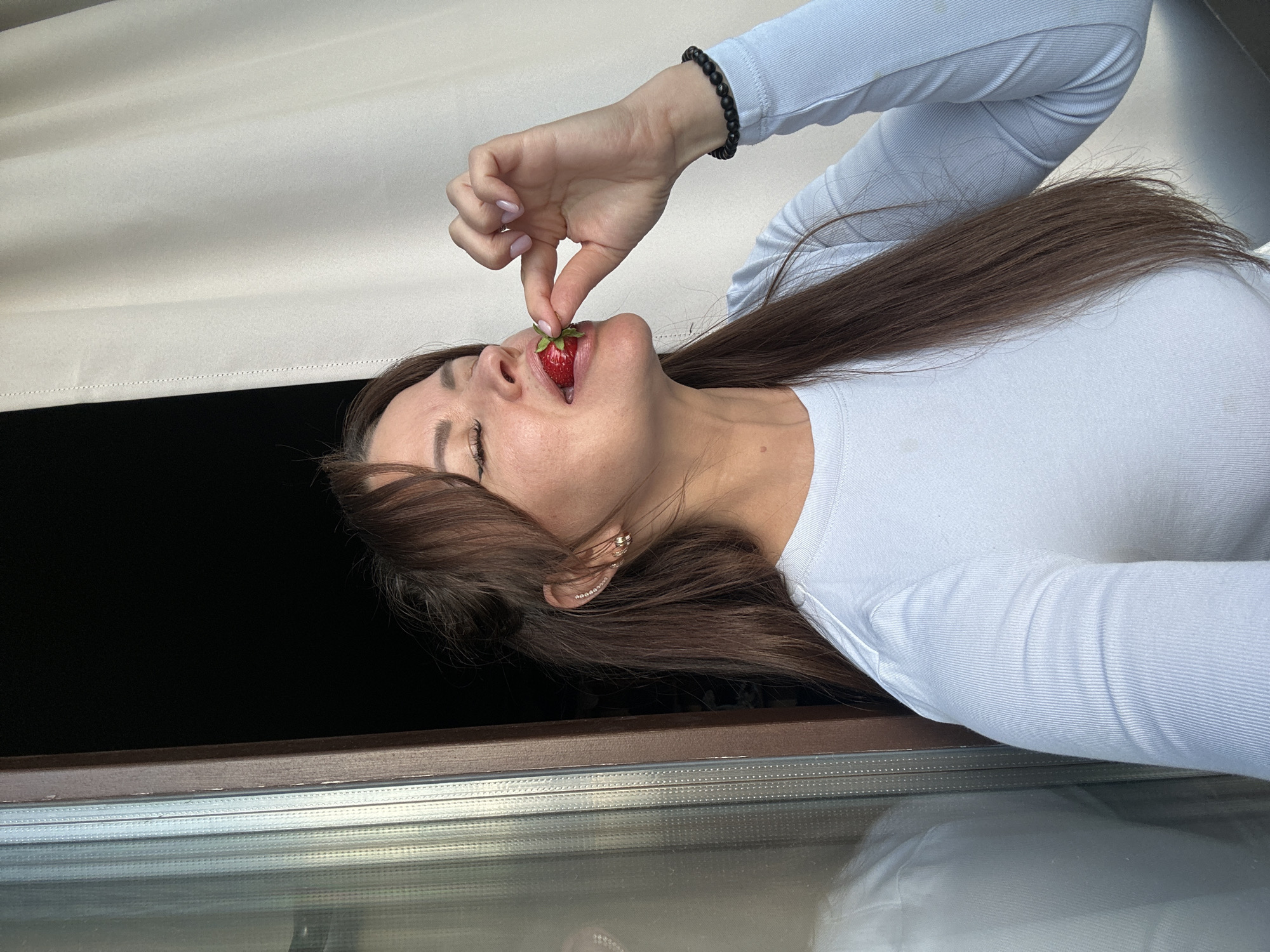 This media file shows a close-up, vertically oriented shot of a young woman with long, brown hair eating a strawberry. She is wearing a white, long-sleeved, ribbed top. Her head is tilted back, and her eyes are closed as she bites into the strawberry, which is held between her fingers. A black beaded bracelet is visible on her right wrist.
The background appears to be an indoor setting with a light-colored blind or curtain partially visible at the top. Below the woman, there's a dark space, possibly an open window or doorway, with a brown frame and a reflective surface below that, suggesting glass. The lighting is soft and natural, indicating it could be daytime. No text is visible in the image.
