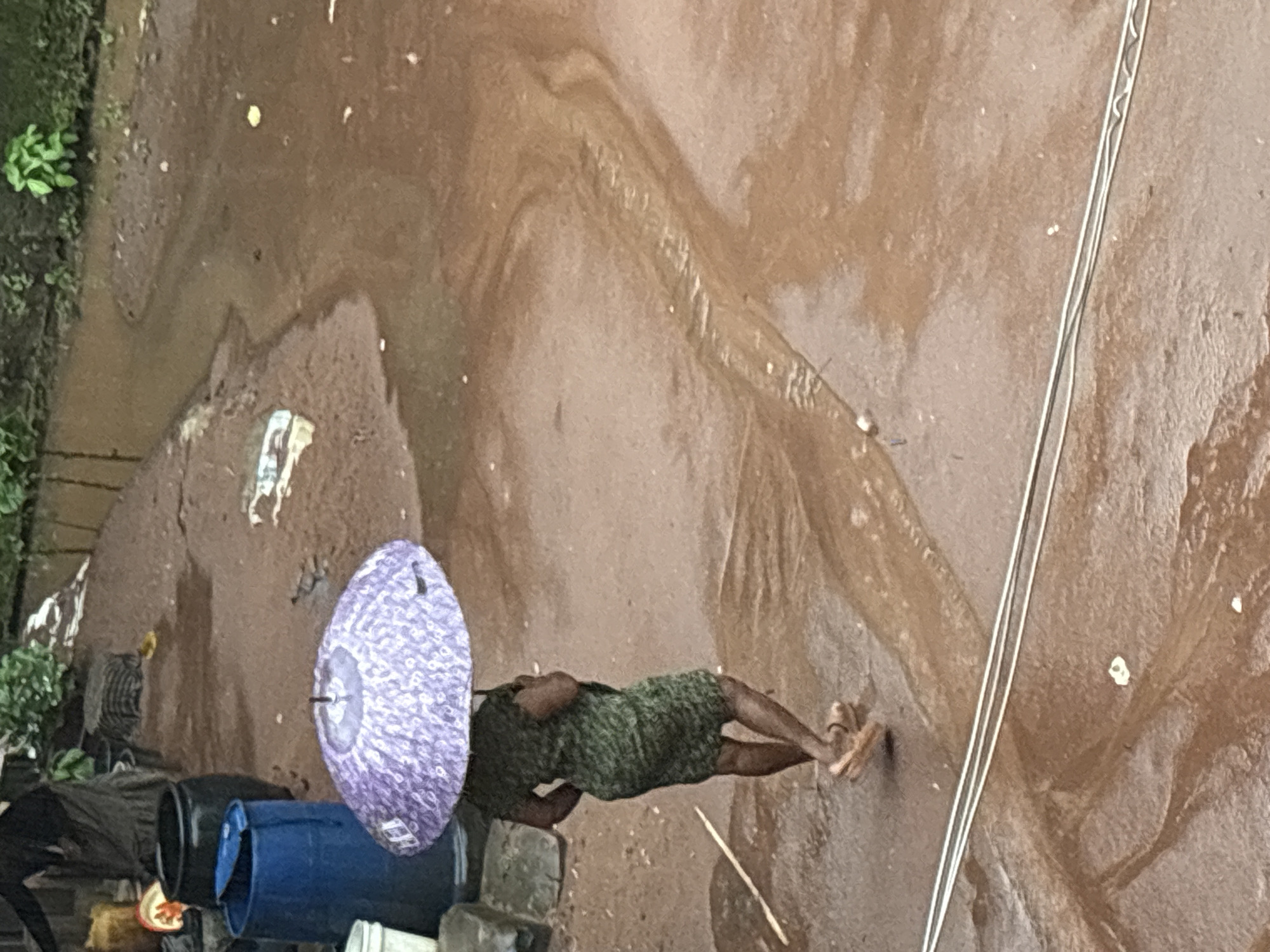 A person walks under a purple umbrella through a muddy, flooded street in Onitsha, Nigeria, during what appears to be a heavy rain. The street is a mix of reddish-brown mud and standing water, with visible ripples and currents indicating the water flow. The person, dressed in a green patterned dress and sandals, is walking away from the camera, shielding themselves from the rain. In the background, several large blue and black barrels are visible, along with some greenery and structures that suggest an urban environment. The overall scene depicts a typical weather condition in the region, characterized by heavy rainfall and its impact on urban infrastructure.