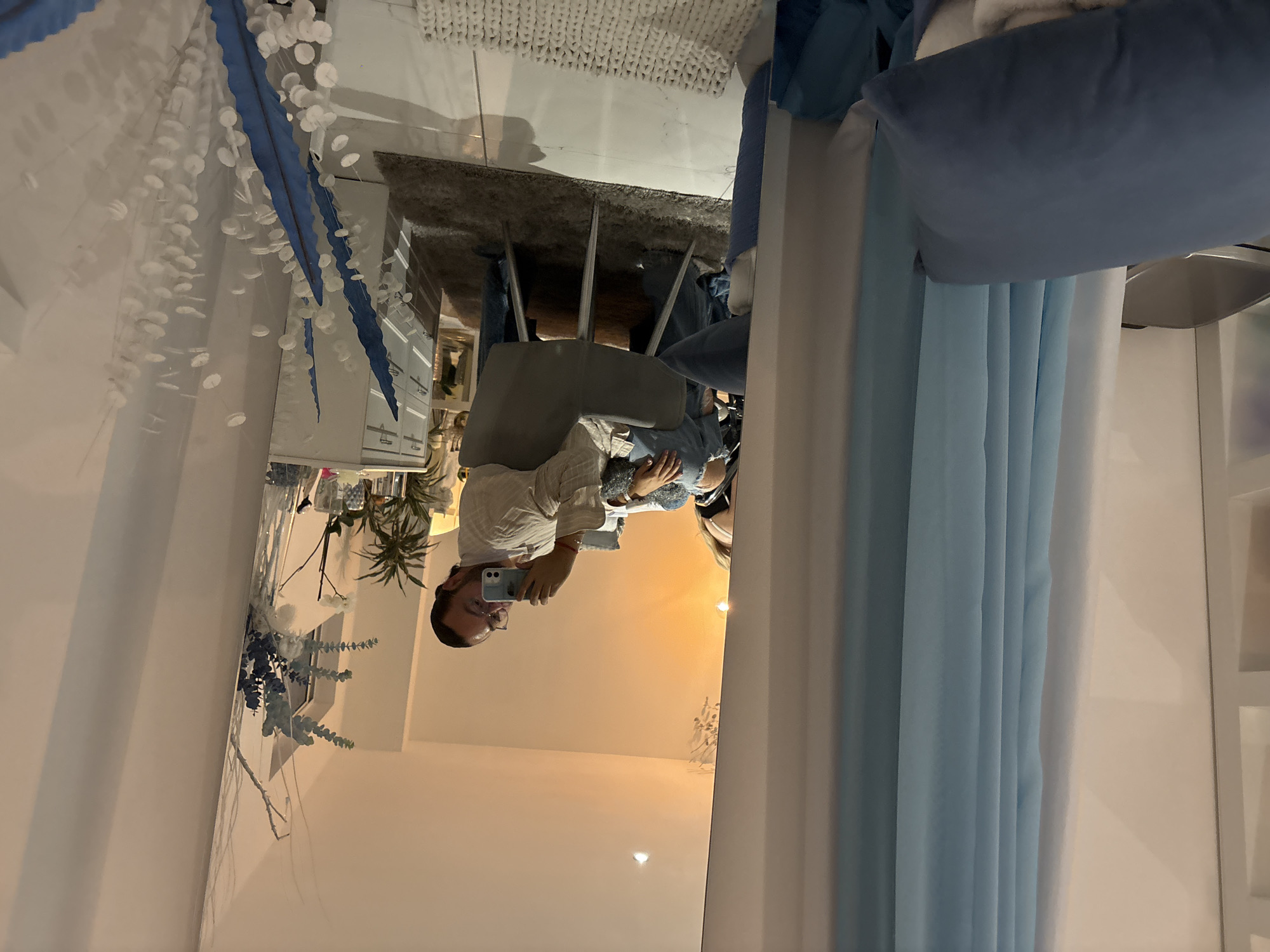 A person is reflected in a mirror, seated on a chair with a light-colored cushion and back, inside a room. The person is holding a cell phone. The room includes a white dresser with decorations, a gray rug on a light-colored floor, and blue curtains. The location in Puebla, Mexico, cannot be determined from the image.