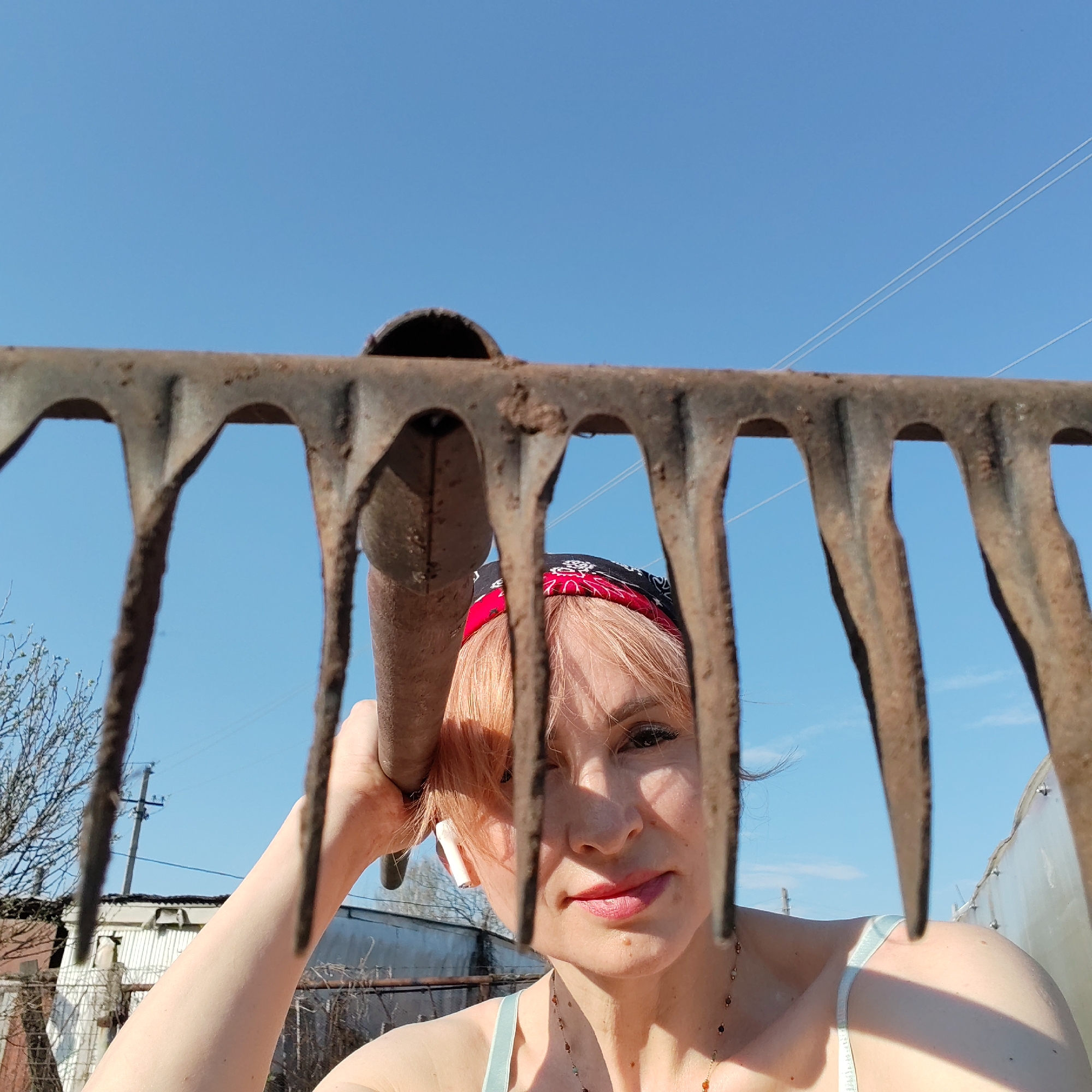 A woman holds a rusty rake up to the camera, framing her face and upper body with the rake's tines and handle. She wears a red and black bandana, has short, light orange hair, and a necklace. A white wireless earbud is visible in her left ear. She is wearing a light-colored top with thin straps. The background includes a clear blue sky, utility poles with wires, and structures that appear to be buildings and greenhouses. The specific location is the city of Chaikovsky, Russia.