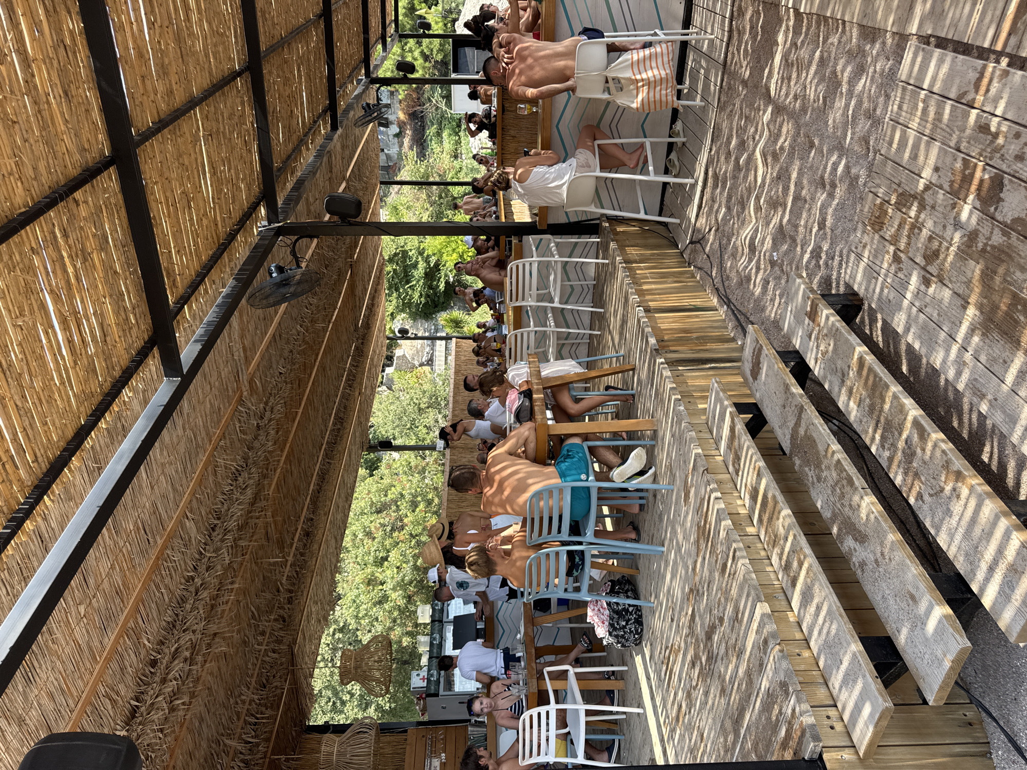 The image depicts a vibrant beachside bar and restaurant scene in Mirište, Montenegro, characterized by a rustic, open-air design with a thatched roof and wooden decking. Numerous individuals are engaged in various activities, including dining at tables, sitting at the bar, and socializing. The environment is distinctly Mediterranean, with lush greenery visible in the background. The overall atmosphere suggests a leisurely and enjoyable day at a popular coastal venue.