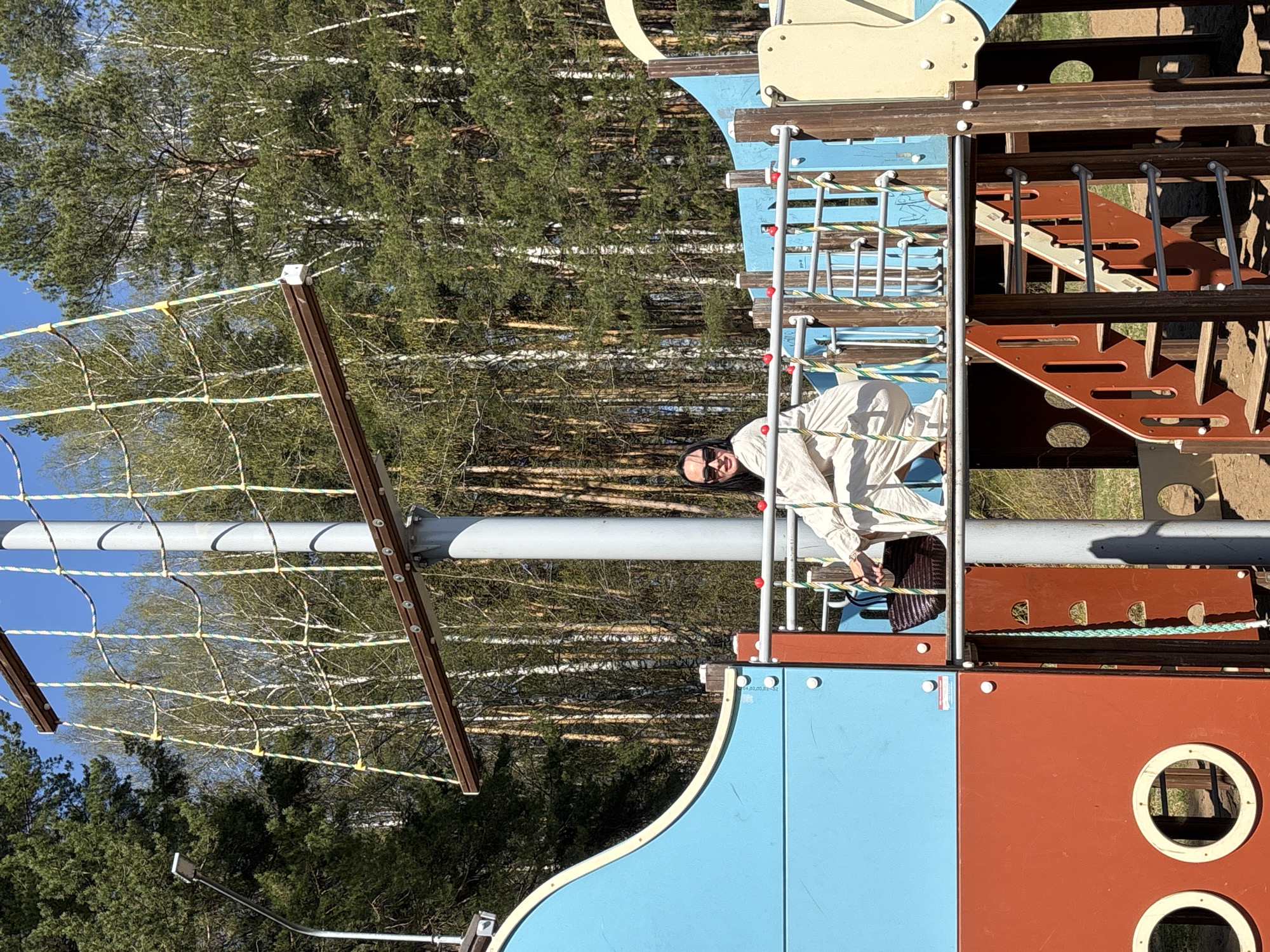 The image depicts a person, apparently an adult female, on a colorful playground structure in an outdoor environment. The structure is constructed of wood and metal, with blue, red, and yellow panels. It includes a climbing net on the left, ladders, and other elements typical of a playground. The woman is wearing a light-colored outfit and sunglasses. In the background, there's a dense forest consisting primarily of birch and pine trees. A street light is visible in the distance. Based on geotagging information, the location is identified as the city of Chaikovsky in Russia. The time of day is likely daytime due to the presence of bright natural light.