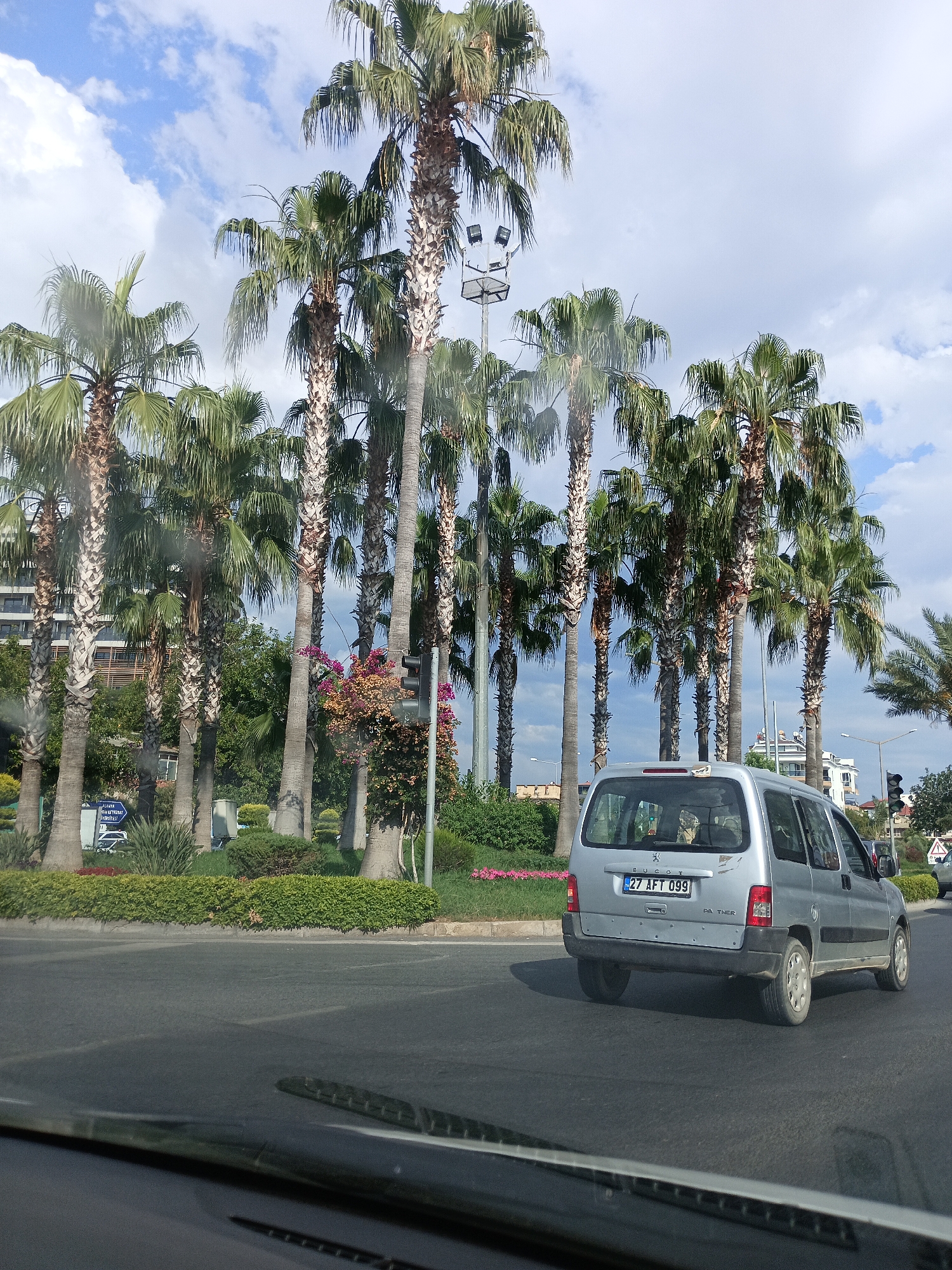 A silver Peugeot Partner van, license plate 27 AFT 099, is traveling on an asphalt road in Alanya, Turkey. The van is viewed from behind and slightly to the right. The road is bordered by a median strip with manicured green bushes and scattered pink flowers. Behind the median strip, a row of tall palm trees lines the street, with a modern building and more palm trees visible in the distance. The sky is partly cloudy with a bright blue sky. A traffic light is visible on the right side of the frame. The image appears to be taken from another vehicle, as evidenced by the dashboard and windshield wipers in the foreground.