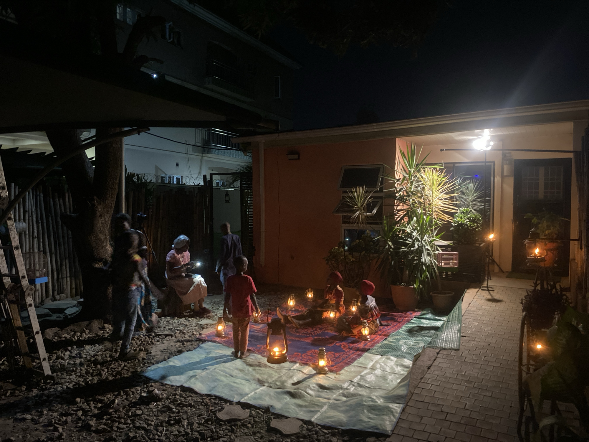 The image depicts an outdoor scene at night in what appears to be the city of Barden Yana, Nigeria. A group of individuals are gathered on a light blue tarp and a red and blue patterned mat that sits on the edge of a stone and brick paved area.
Several people are standing, sitting, and interacting around the mats. A child in a red shirt stands near a lantern and faces the seated group. Others are standing to the left, and one appears to be using a cell phone. The group is lit by multiple kerosene lanterns placed on and around the mats.
In the background, there is a one-story building with an orange exterior and an attached two-story building. Plants are in pots along the building. A light shines from the covered porch. There is also a tree and bamboo fencing on the left side of the image. A ladder leans against the bamboo fence. Stone aggregate ground cover surrounds the tarp area on the left. There are multiple kerosene torch lights along the brick pathway.
