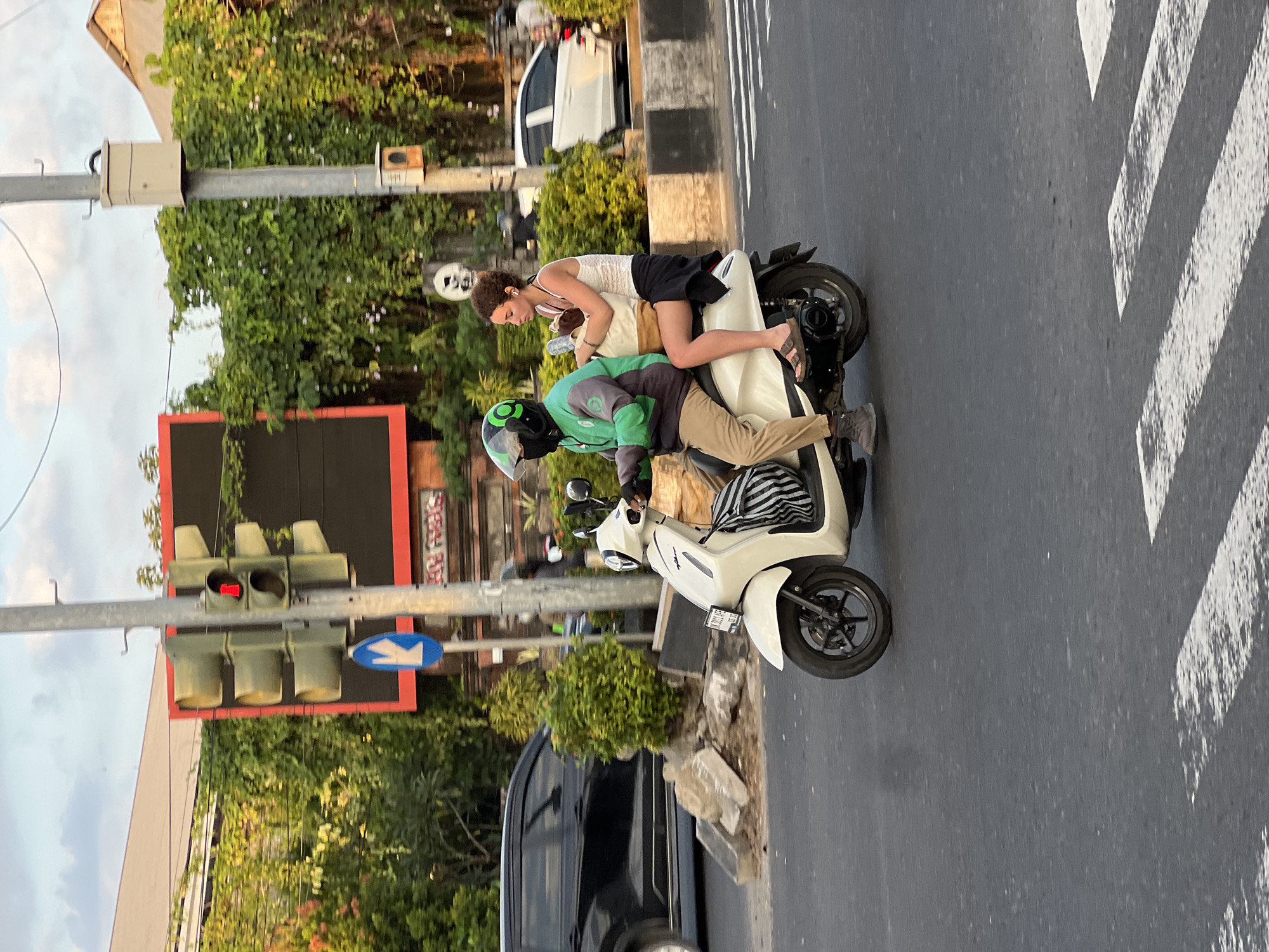 The image captures a street scene in Denpasar Barat, Indonesia, under a cloudy sky. Two individuals are riding a white scooter on a paved road. The person in front, wearing a green jacket with a black collar and cuffs, and a black helmet with a green logo, is driving. They are also wearing tan pants and dark shoes. The passenger behind, a young woman with curly hair, wears a white tank top and a black skirt. She is holding a large light-colored tote bag and has a water bottle. The scooter has a license plate with the characters "BK 3001 BFE".
In the background, a black car is visible on the left, and a white car is further down the road on the right. Traffic lights are mounted on a metal pole, with the red light illuminated. A blue directional arrow sign is also present on the pole. Lush green foliage and trees cover the area behind the road, with a large billboard framed in red to the left of the traffic lights. The road surface shows white pedestrian crossing lines. The overall impression is of a typical urban setting during daylight hours.