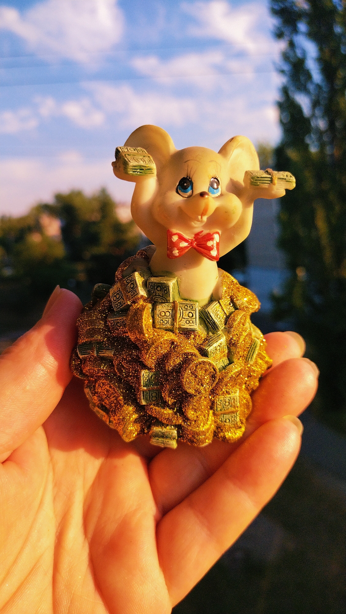 An overhead, medium shot captures a human hand holding a figurine of a smiling mouse. The mouse is made of a light-colored material, with blue eyes, a red and white polka dot bowtie, and is adorned with stacks of money. It is standing on a pile of what appears to be gold coins and more stacks of money, all of which are dusted with glitter. The mouse is held in the palm of a hand with visible lines and a clean fingernail. The background is blurred, showing trees and a sky with clouds, indicating an outdoor setting. The lighting suggests it is either sunrise or sunset, casting a warm glow on the scene. This image was taken in Horishni Plavni, Ukraine.