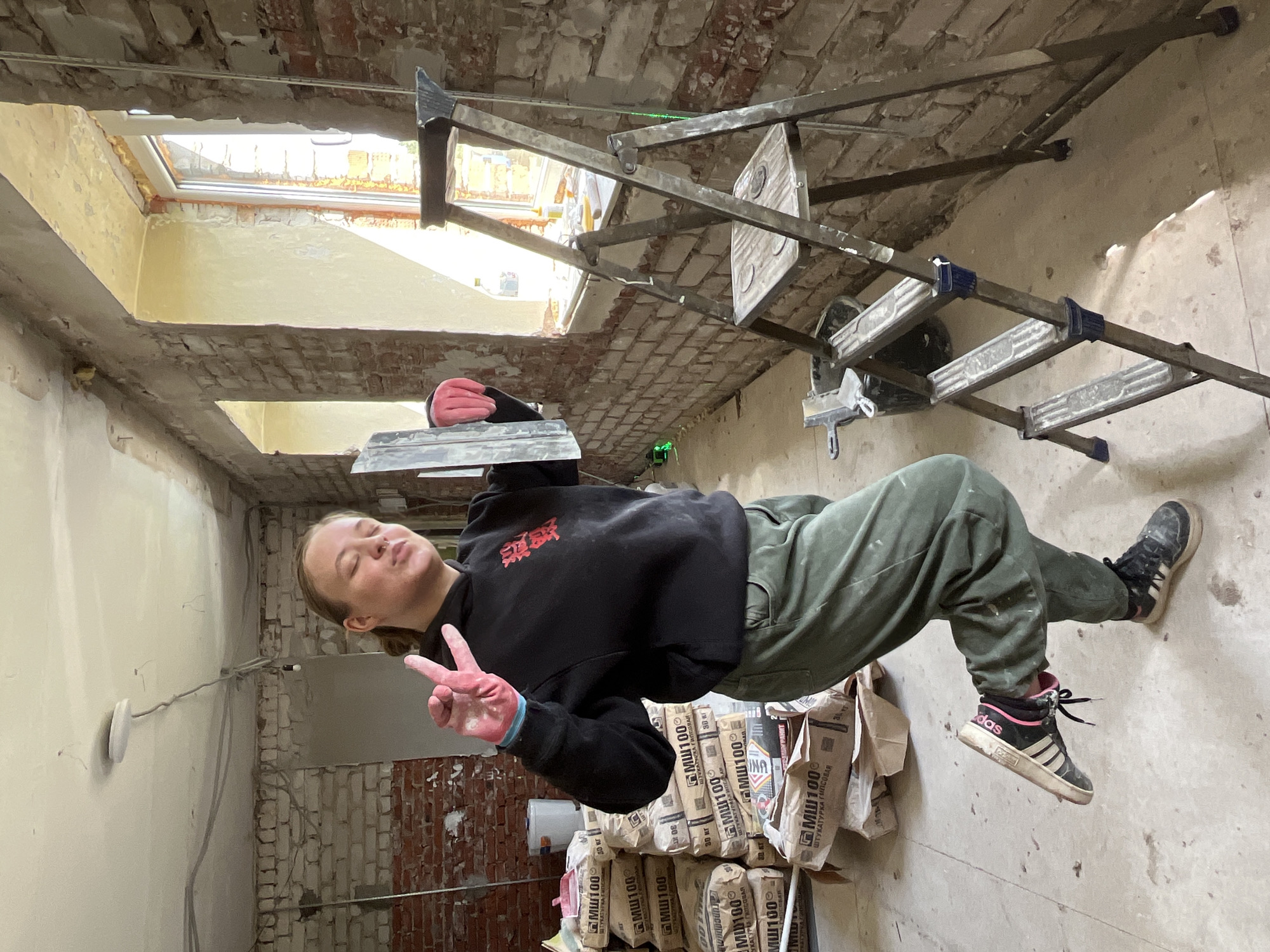 A woman wearing a black sweatshirt, green pants, and Adidas sneakers is standing in a room under construction. She is giving a peace sign with her left hand and holding a trowel in her right hand. In the background, there are stacks of bags of MШ100 plaster from "ТРАМОГРАНИТ" in the city of Фоки, Russia, and a ladder is visible. The walls are partially brick and partially plastered. There are also skylights in the ceiling.