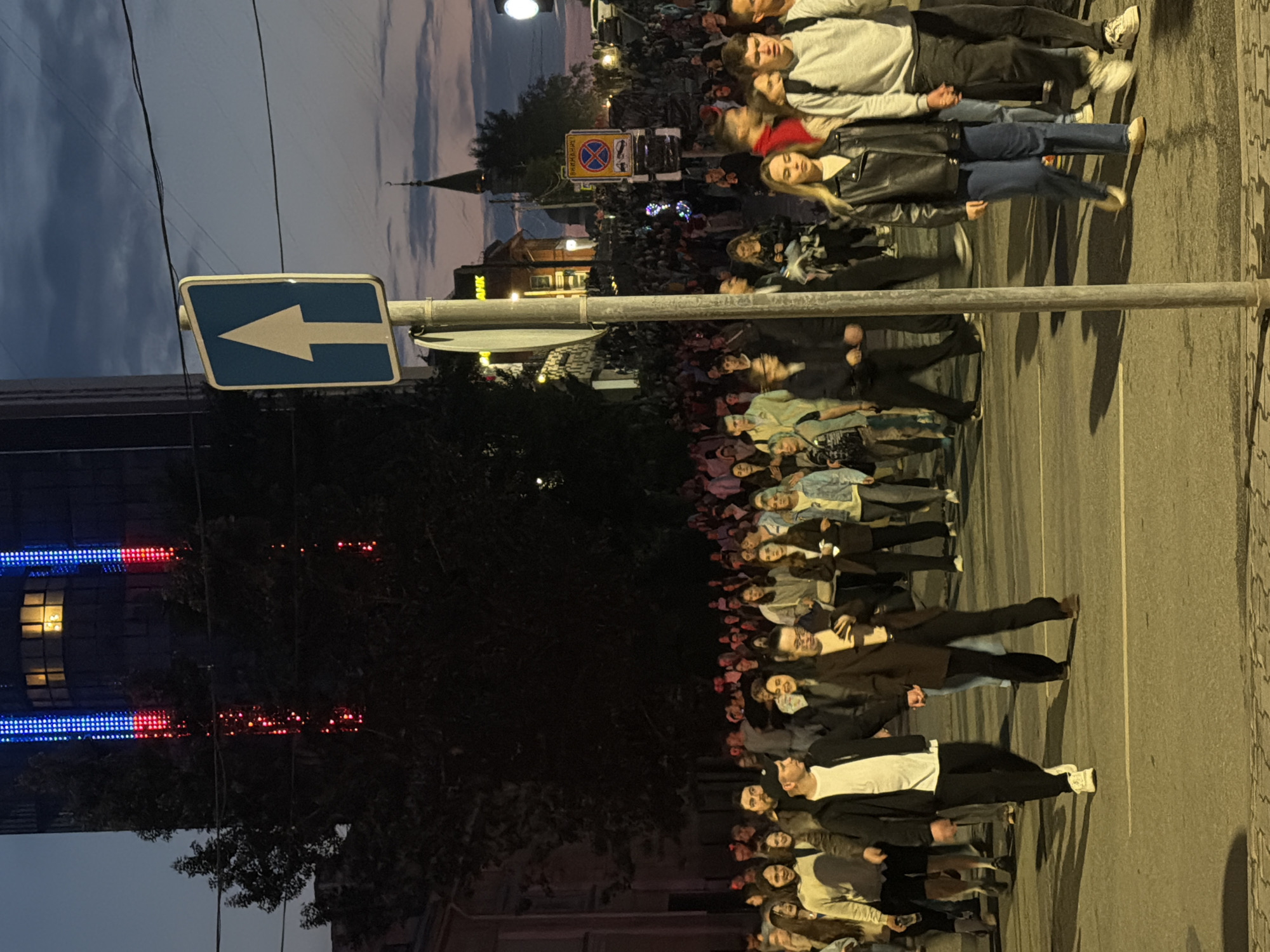 A crowd of people are walking down a street at dusk. The street appears to be paved and has white lane markings. A tall, modern building with blue and red lights is visible in the background, along with a church steeple. There are street signs and power lines visible. The people are dressed in casual clothing and some are holding hands. The lighting suggests it is nighttime or late evening. The scene appears to be a public gathering or event. The location is the city of Perm, Russia.