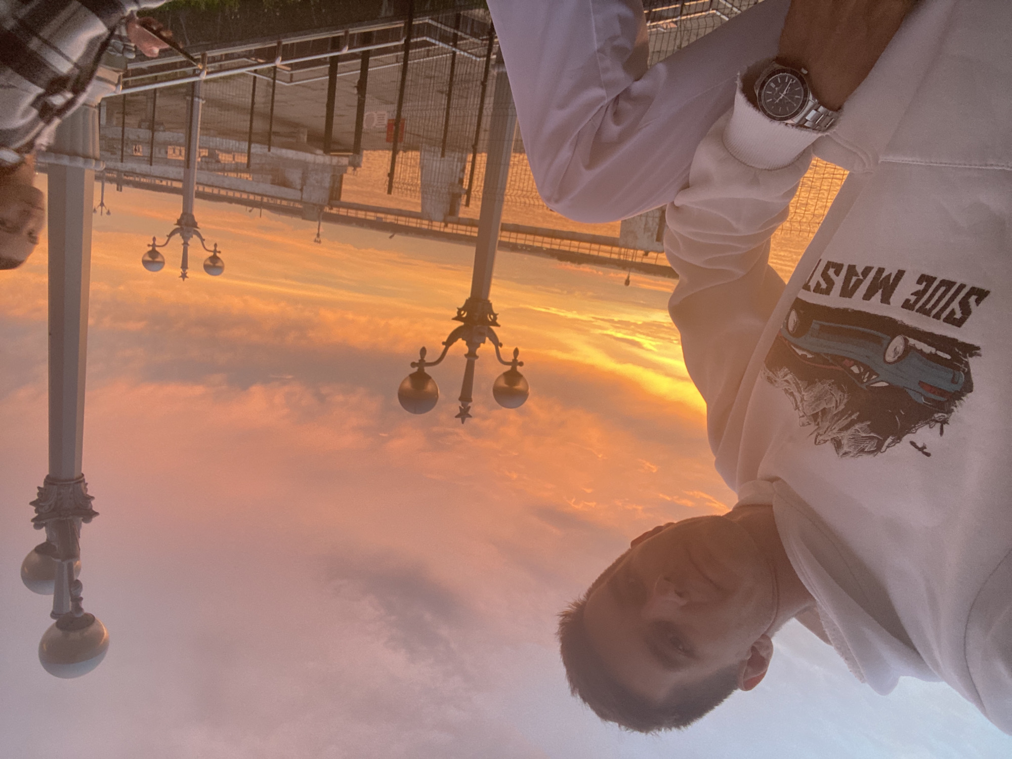 A man wearing a white hoodie with a graphic of a blue sports car and mountains, along with the text "SIDE MAST" and white pants, is posing for a selfie during sunset. He is wearing a silver wristwatch. To his right, a woman in a plaid shirt is seated and looking at her phone. The background features a river, a bridge, and streetlights, illuminated by the warm orange and pink hues of the sunset. The location is likely on the embankment in Novokuybyshevsk, Russia, given the visual elements.