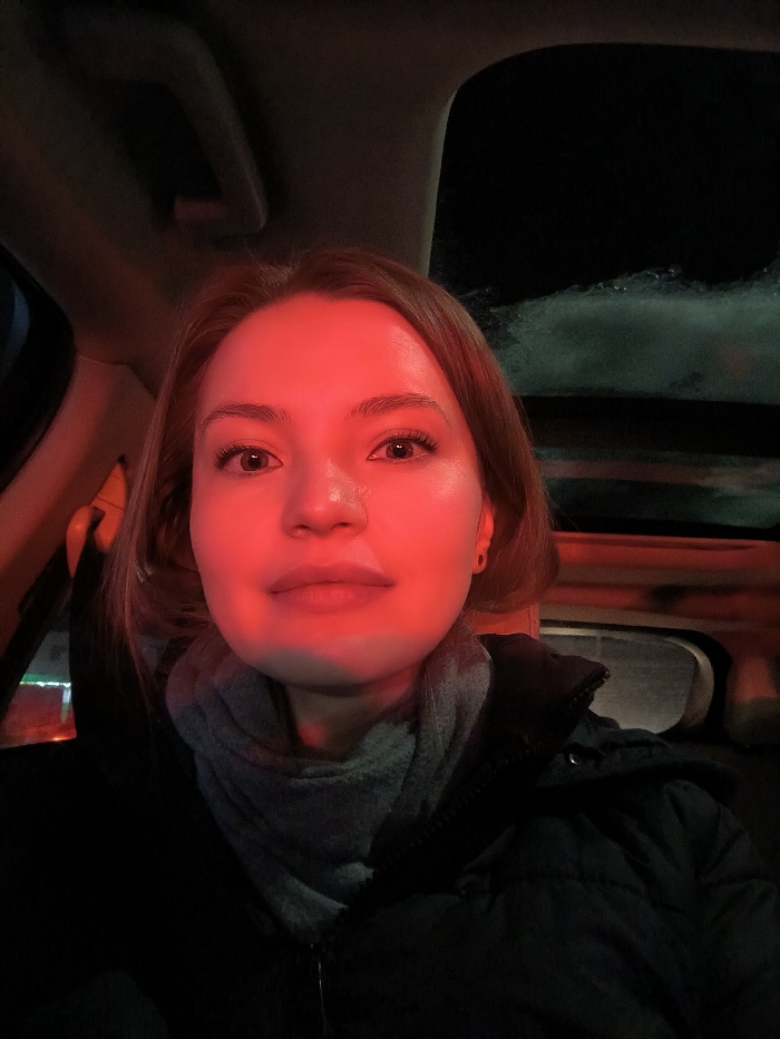 A selfie shows a fair-skinned woman with reddish-brown hair inside a car at night. Her face is illuminated by a strong red light. She's wearing a dark puffer jacket and a light grey scarf. A black car interior with the car roof above and the side window to the left is visible. The window shows outside darkness, with a hint of white reflecting off of the dark background. The red lighting casts strong shadows and creates a dramatic effect.