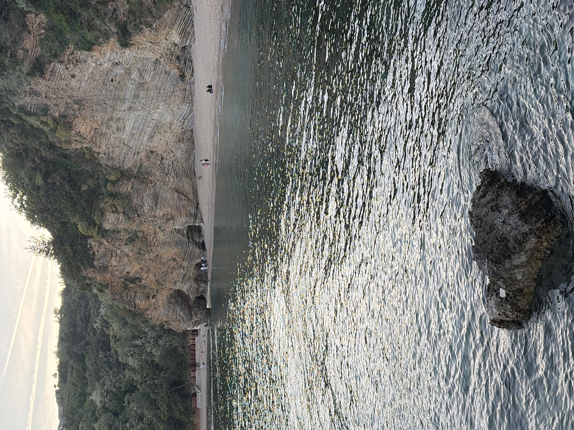This vertical image captures a tranquil coastal scene in Budva, Montenegro, featuring a steep rock face, a sandy beach, and the sparkling sea under bright daylight.
On the left, a towering, layered rock cliff dominates the landscape, exhibiting clear geological stratification and adorned with patches of green vegetation and trees. The intricate patterns of the rock layers are a prominent feature.
Below the cliff, a narrow strip of light-colored sandy beach extends along the coastline. Several small figures, presumably people, are scattered on the beach. In the mid-ground, a pair of individuals can be seen closer to the water's edge, while further down the beach, faint outlines suggest more people and possibly beach furniture like sunbeds or chairs.
The right side of the image is primarily occupied by the sea, which glitters intensely with reflections from the sun, creating a dynamic pattern of light on the water's surface. In the lower right foreground, a solitary dark rock rises from the water, causing small ripples around its base. The sunlight suggests it is either morning or late afternoon. The sky, visible in the upper left, appears clear. There is no visible text in the image.