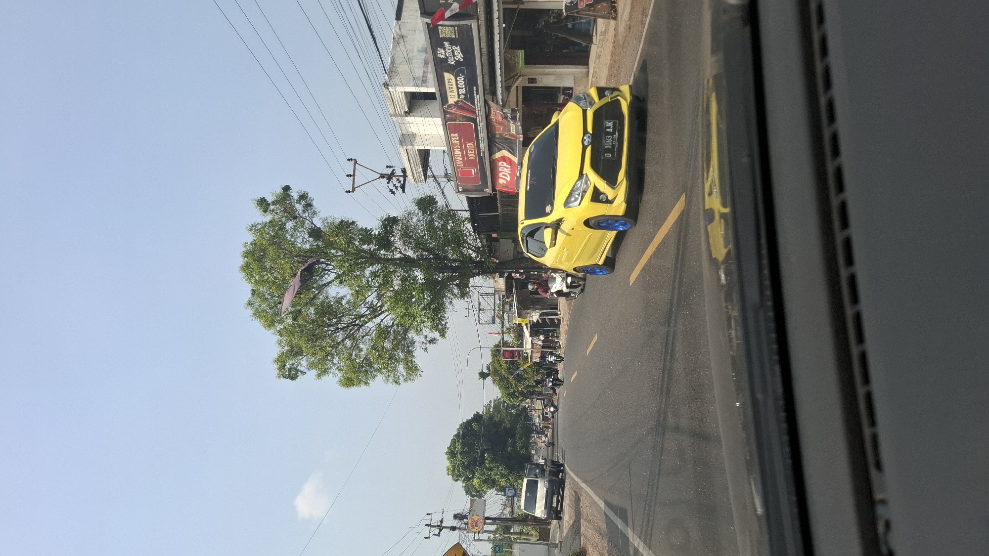 The image depicts a street scene in Cikaroya, Indonesia, under a clear blue sky. A bright yellow car with blue rims is prominently featured in the foreground, driving on a paved road. The road has yellow lane markings. To the left of the road, a white van is parked. Further down the road, several motorcycles are visible, with riders on them. On the right side of the road, there are buildings with storefronts, some displaying advertisements. A large green tree with sparse foliage stands in the middle ground, and a purple banner or flag is attached to one of its branches. Utility poles and electrical wires are present throughout the scene, indicating an urban or semi-urban environment. The perspective suggests the photo was taken from inside another vehicle.