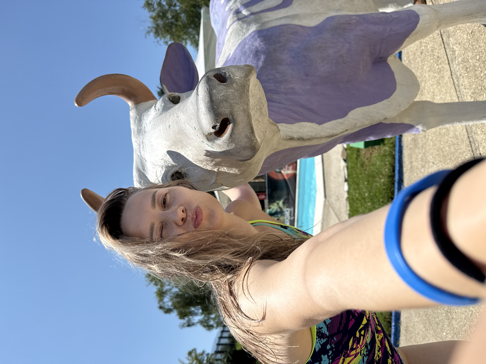 In the city of Nebug, Russia, a woman with wet blonde hair and a playful expression winks at the camera while taking a selfie. She is wearing a colorful swimsuit and a blue wristband. Beside her stands a large, white cow statue with purple markings. The background shows a swimming pool with blue water, lush green grass, and trees under a clear blue sky. The scene appears to be an outdoor recreational area, possibly a resort or a water park.