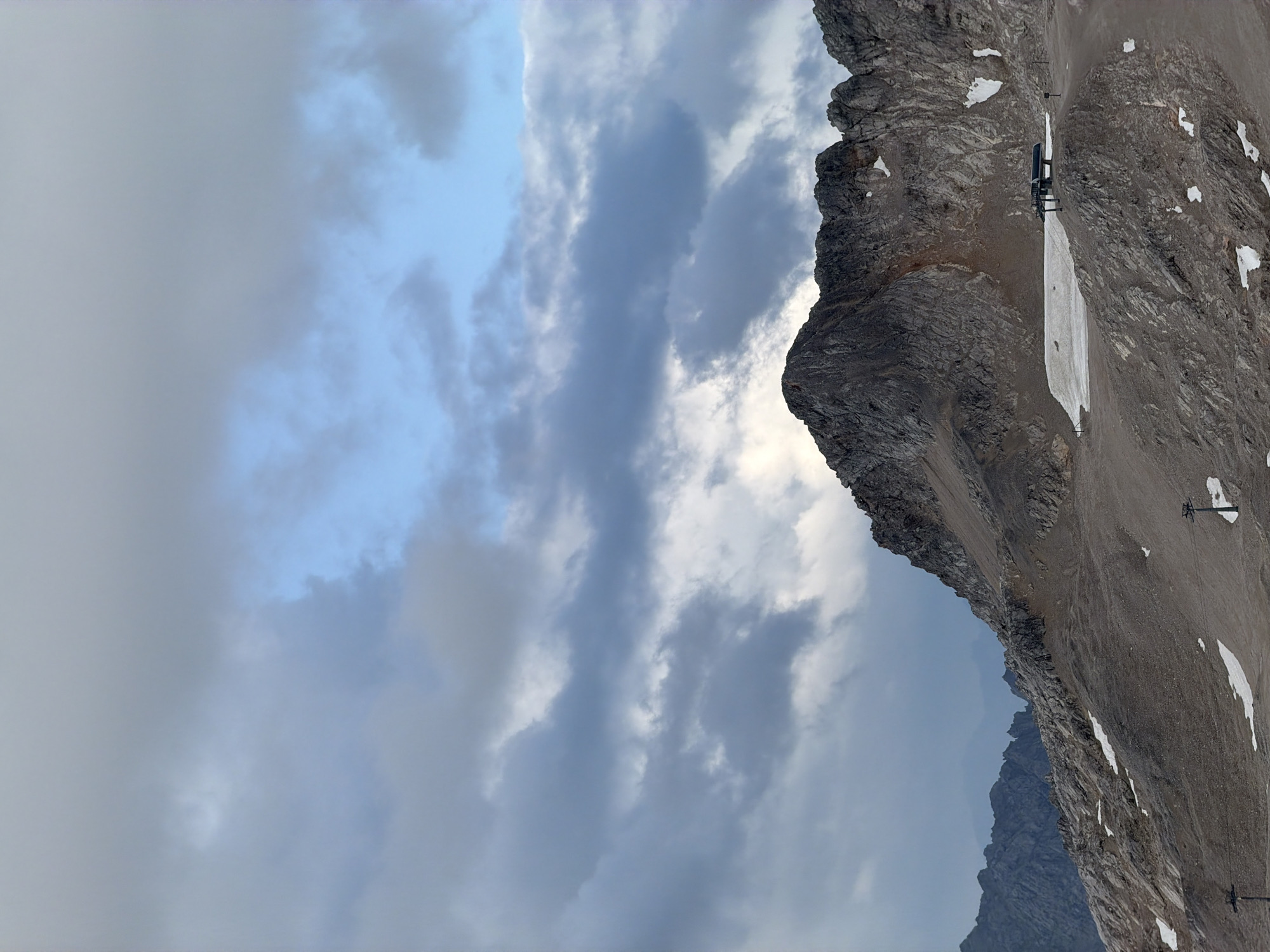 The image depicts a mountainous landscape with a dramatic sky filled with clouds. In the foreground, a rocky, barren slope leads up to rugged mountain peaks. There are patches of snow scattered across the terrain, suggesting a high altitude. Two ski lift towers are visible on the slope. In the mid-ground, a ski lift station is situated on a rocky outcrop, with a small area of snow nearby. The background features more distant mountain ranges, fading into a hazy atmosphere. The lighting indicates it is likely late afternoon or early morning, with a mix of sunlight breaking through the clouds. This scene is likely the Zugspitze, Germany's highest peak, located near Garmisch-Partenkirchen. There are no people or animals visible in the image.