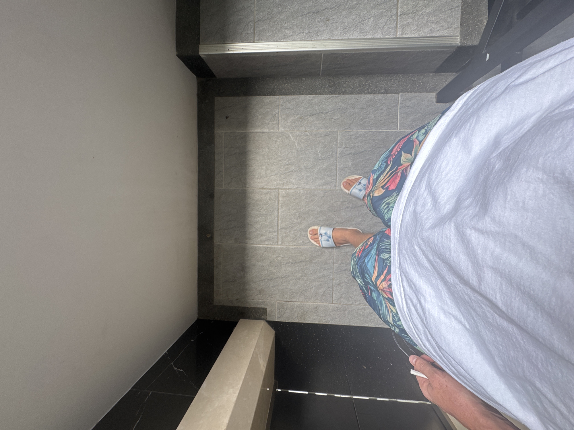 The image shows a first-person perspective of someone standing on a tiled floor, possibly in a bathroom or outdoor shower area. The floor is made of large, grey square tiles. The person is wearing floral-patterned shorts and white slide sandals. Their left arm, holding a white object, is visible in the lower left corner. The background consists of dark tiled walls and a lighter-colored ledge or counter. The lighting suggests an indoor or partially shaded outdoor environment. The scene is devoid of any specific landmarks that would definitively place it in Rawai, Thailand.