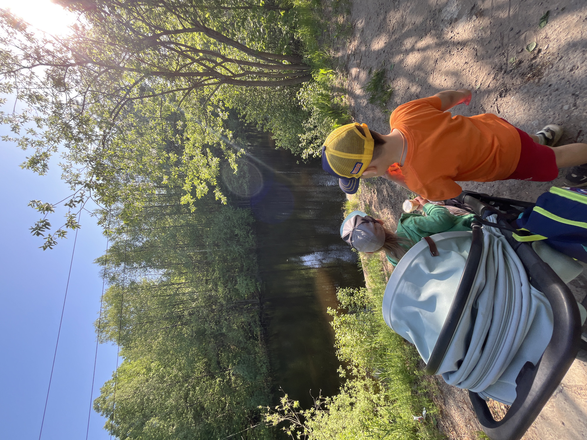 The image shows a dirt path alongside a body of water, likely a canal or narrow river. A young boy in an orange shirt, red shorts, and a yellow baseball cap is standing on the path. An adult is sitting next to a baby stroller. The vegetation consists of trees and shrubs on both sides of the water, creating a wooded area. The photo was taken in Chaikovsky, Russia, given the location information. Overhead power lines are visible against a clear blue sky. The lighting suggests it is a sunny day.