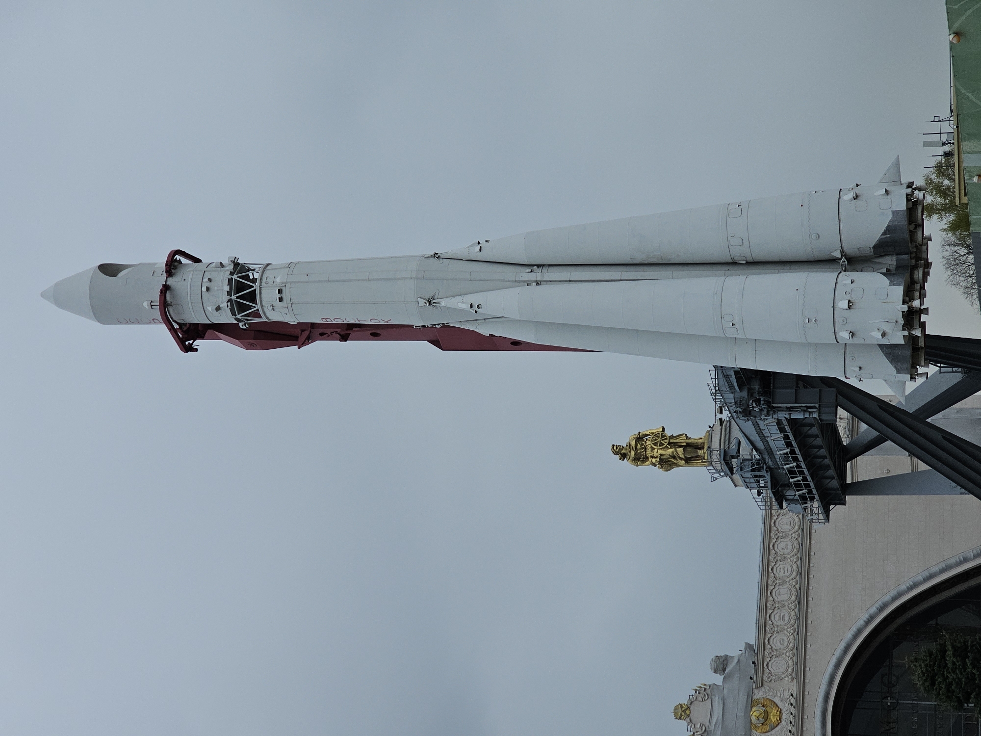 The image displays a large, multi-stage Vostok rocket erected vertically outdoors under an overcast sky. The rocket is predominantly white with red structural elements and lettering. The word "BOCTOK" (Vostok), meaning "East" in Russian, is clearly visible in red text on the side of the rocket's main body.
Adjacent to the rocket and slightly lower, a prominent golden statue stands on an ornate pedestal. This statue depicts a figure, likely a worker or farmer, holding an object that resembles a sheaf or emblem. The statue is positioned in front of a grand building featuring classical Soviet-era architecture, characterized by decorative friezes, detailed pilasters, and a large, arched entrance. The building's facade includes indistinct ornamental crests or symbols.
The rocket is supported by a dark-colored, robust gantry structure. In the background, particularly visible in the upper right of the original image, there are hints of green foliage and a green structure, suggesting a park-like environment. The overall scene depicts a static exhibit, commemorating Soviet space achievements. The uniformly light gray sky indicates an overcast day. No specific activity beyond public viewing of the display is evident. The setting is consistent with a historical or technological exhibition park, such as VDNKh in Moscow, Russia.